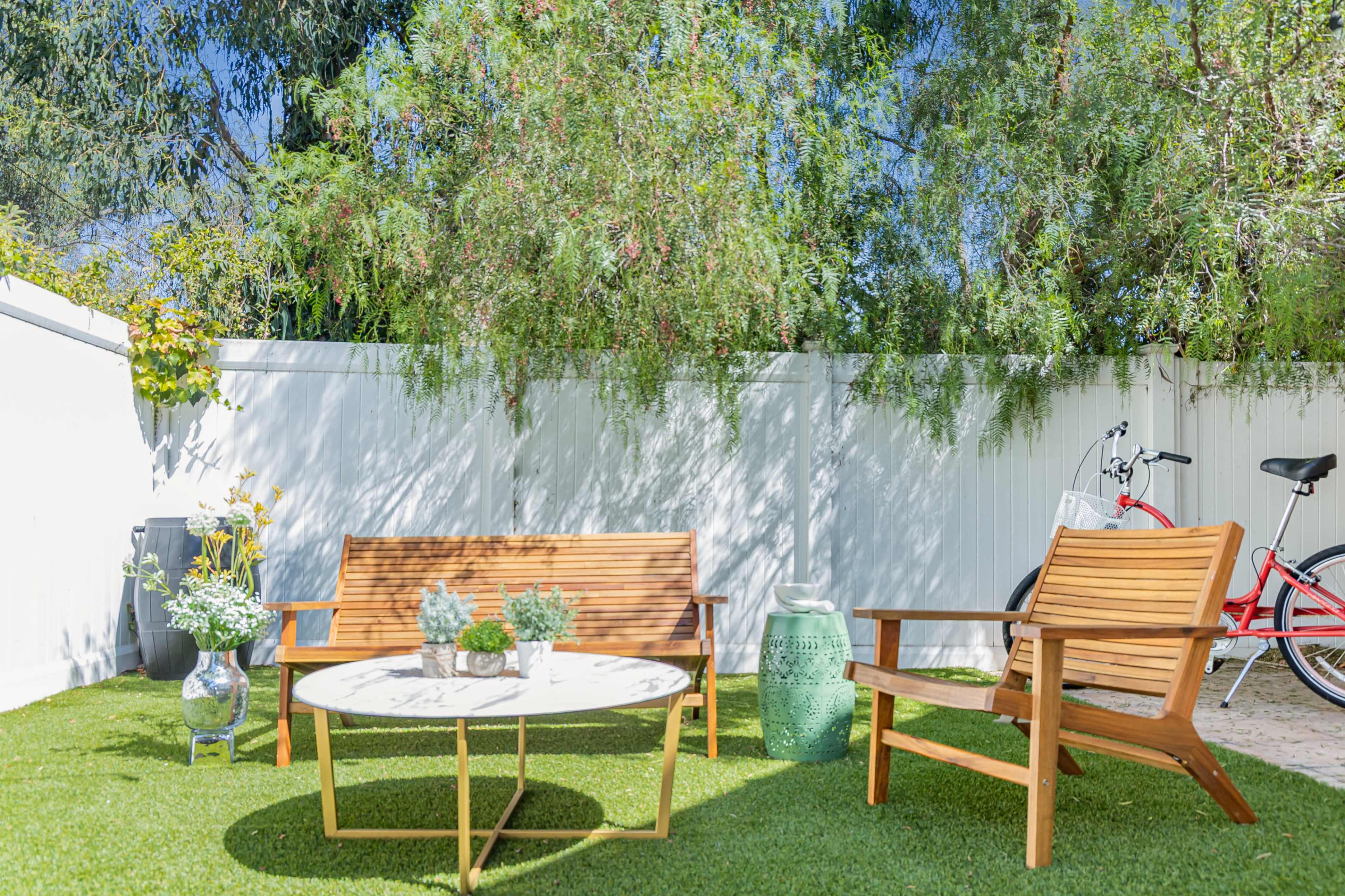 The image shows a sunny outdoor seating area with two wooden chairs, a round table, potted plants, and a bicycle leaning against a fence.