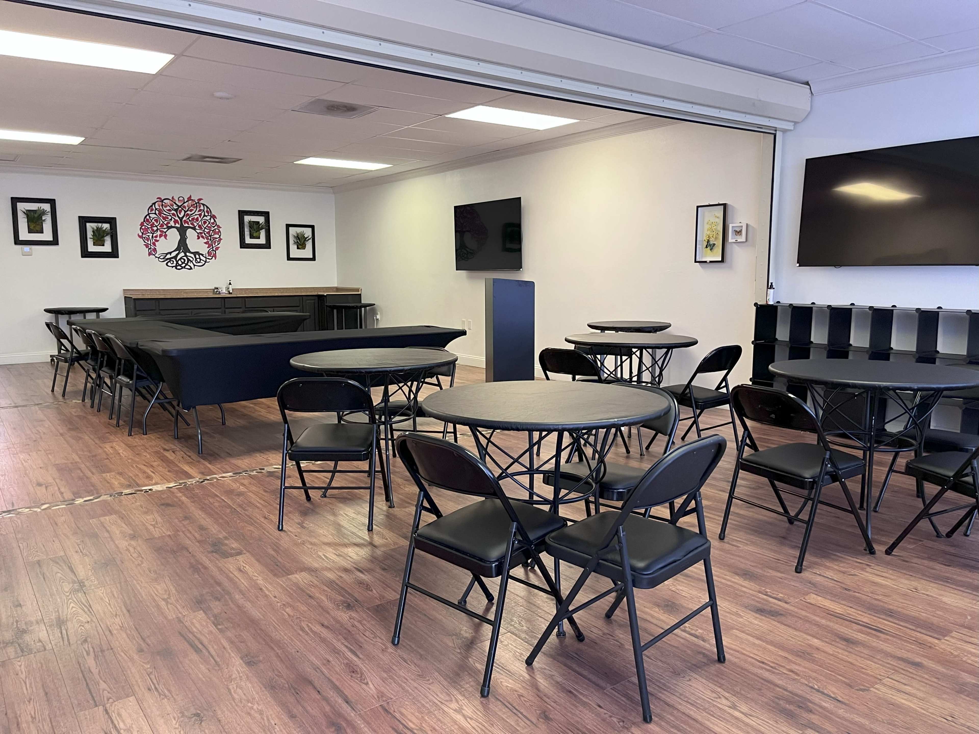 The image shows a spacious room setup with multiple round tables and chairs, featuring a bar area and wall-mounted screens.