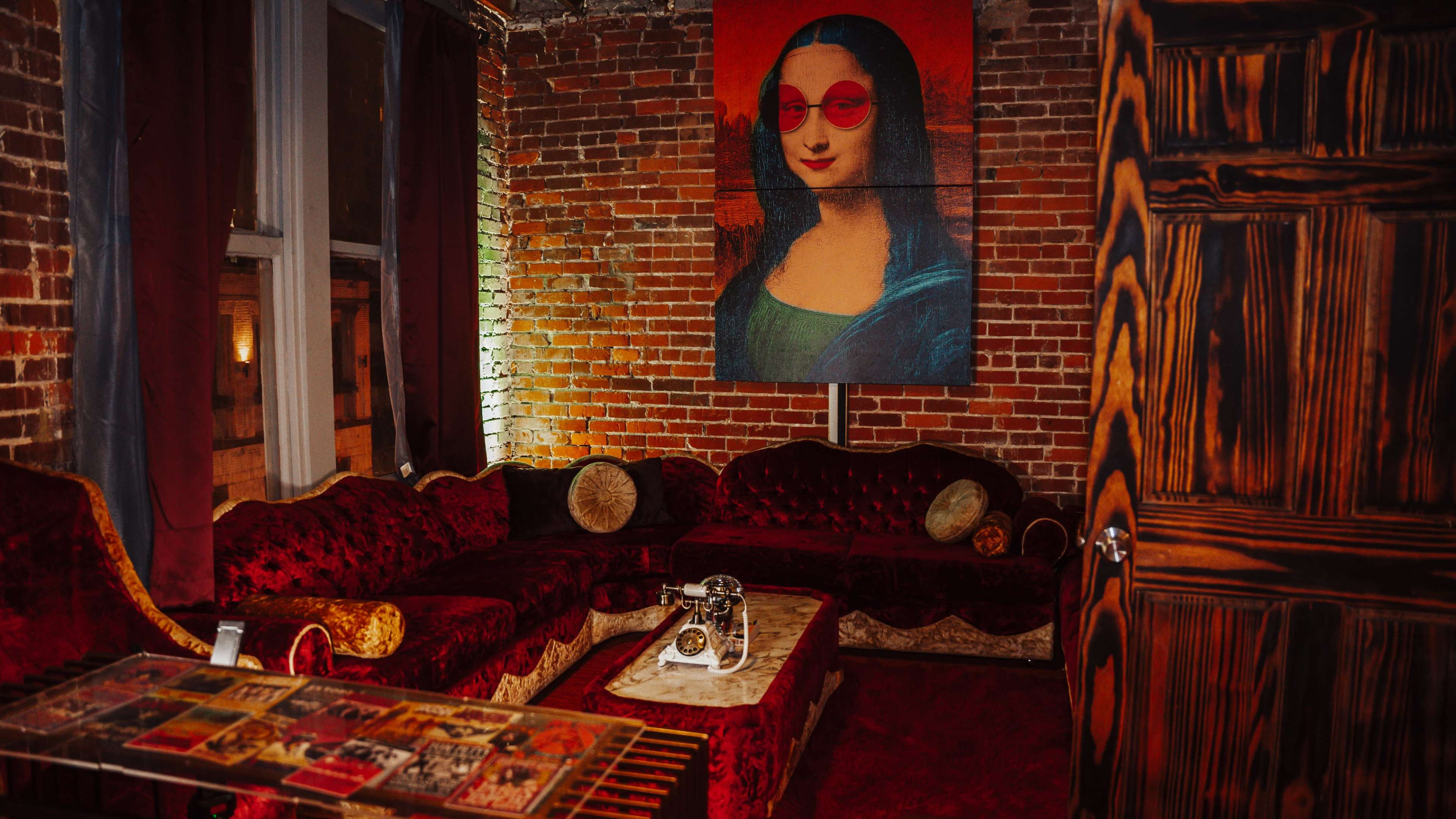 The image shows a stylishly decorated room featuring a red brick wall, a plush red couch, a coffee table with magazines, and a large painting of a woman with sunglasses on the wall.