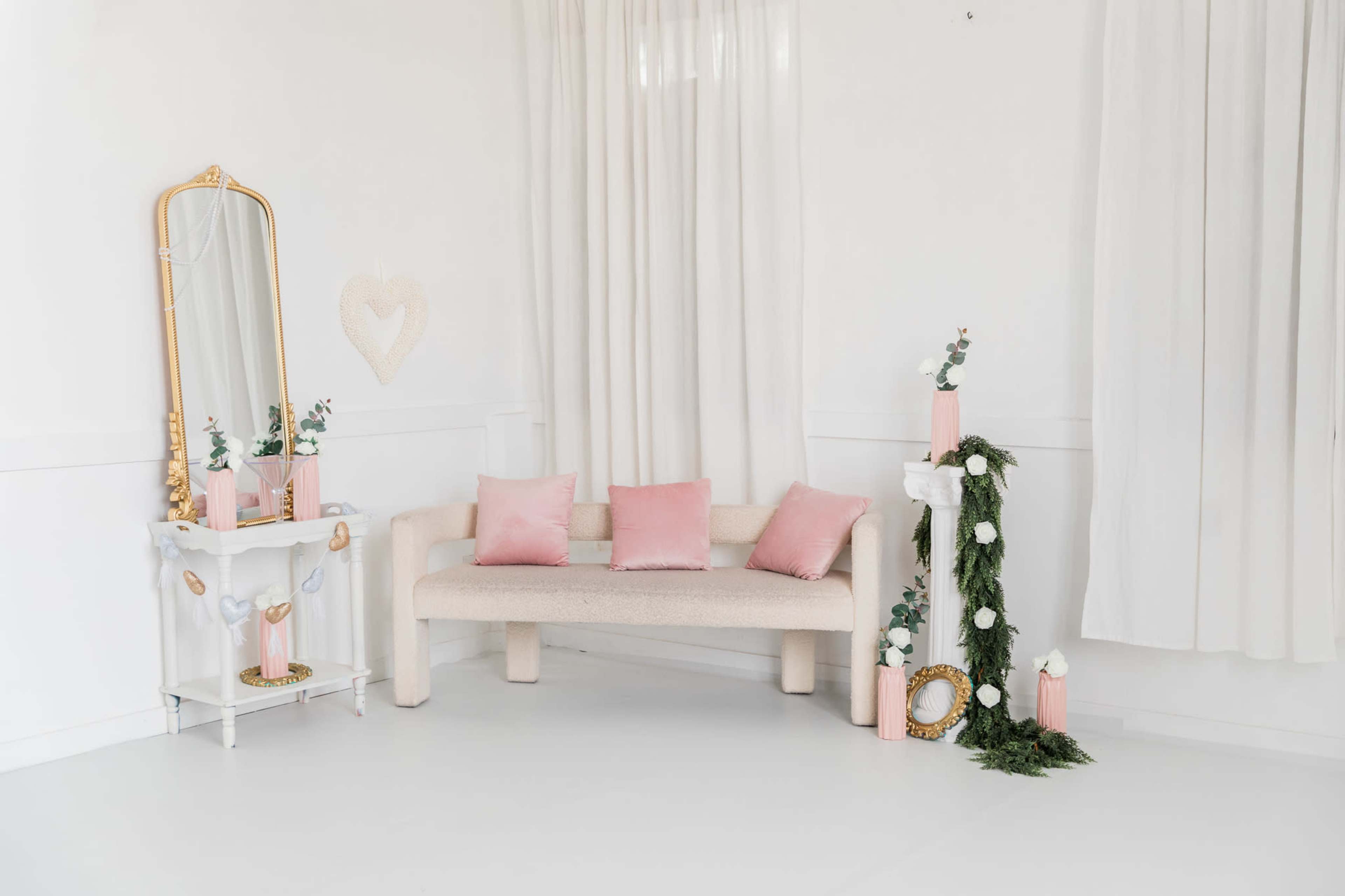 The image features a modern, minimalist living space with a light-colored sofa, pink accent pillows, a decorative mirror, and greenery in vases.