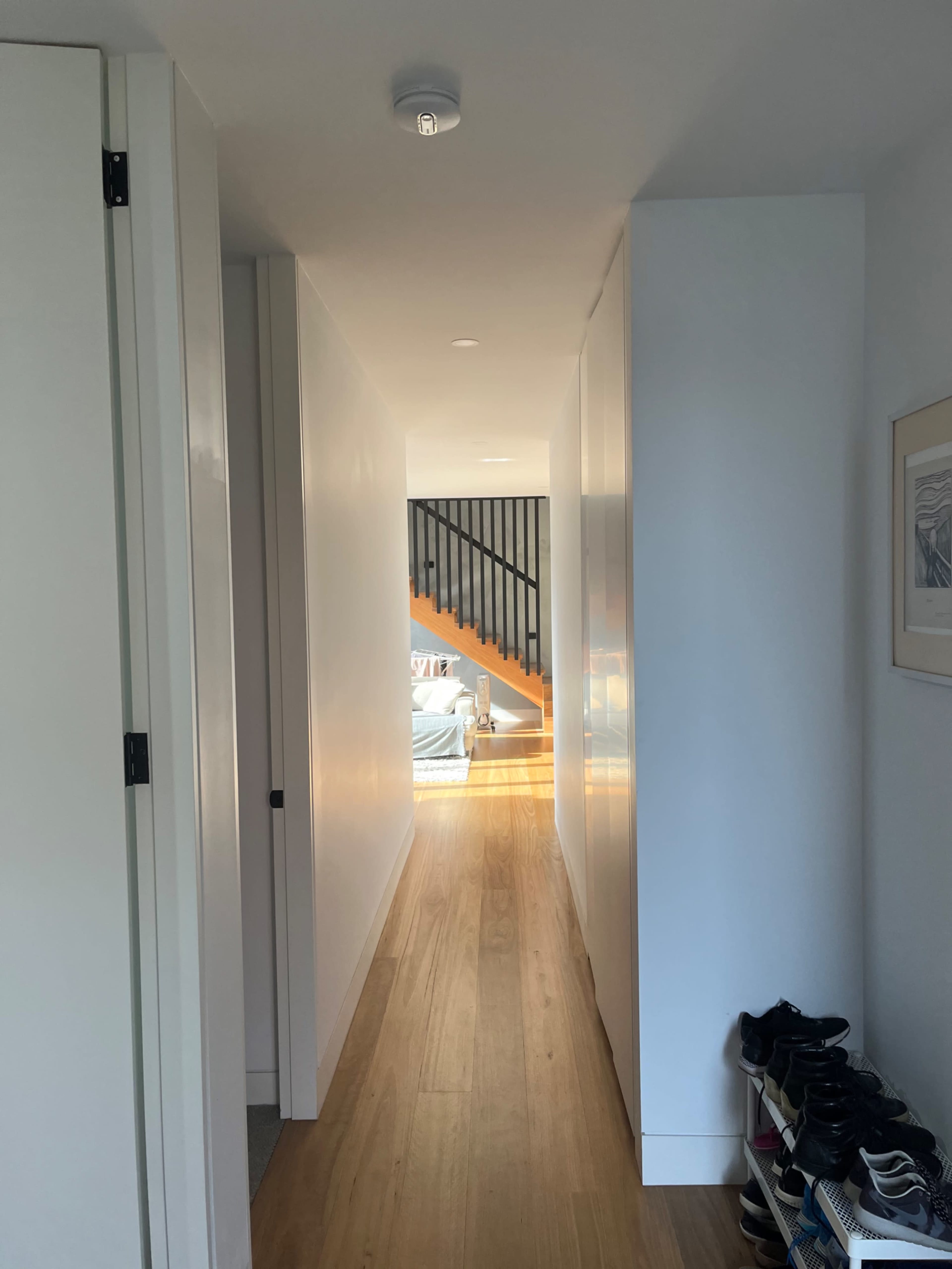 A narrow hallway with wooden flooring leads to a bright area at the end, flanked by closed doors and a shoe rack on one side.