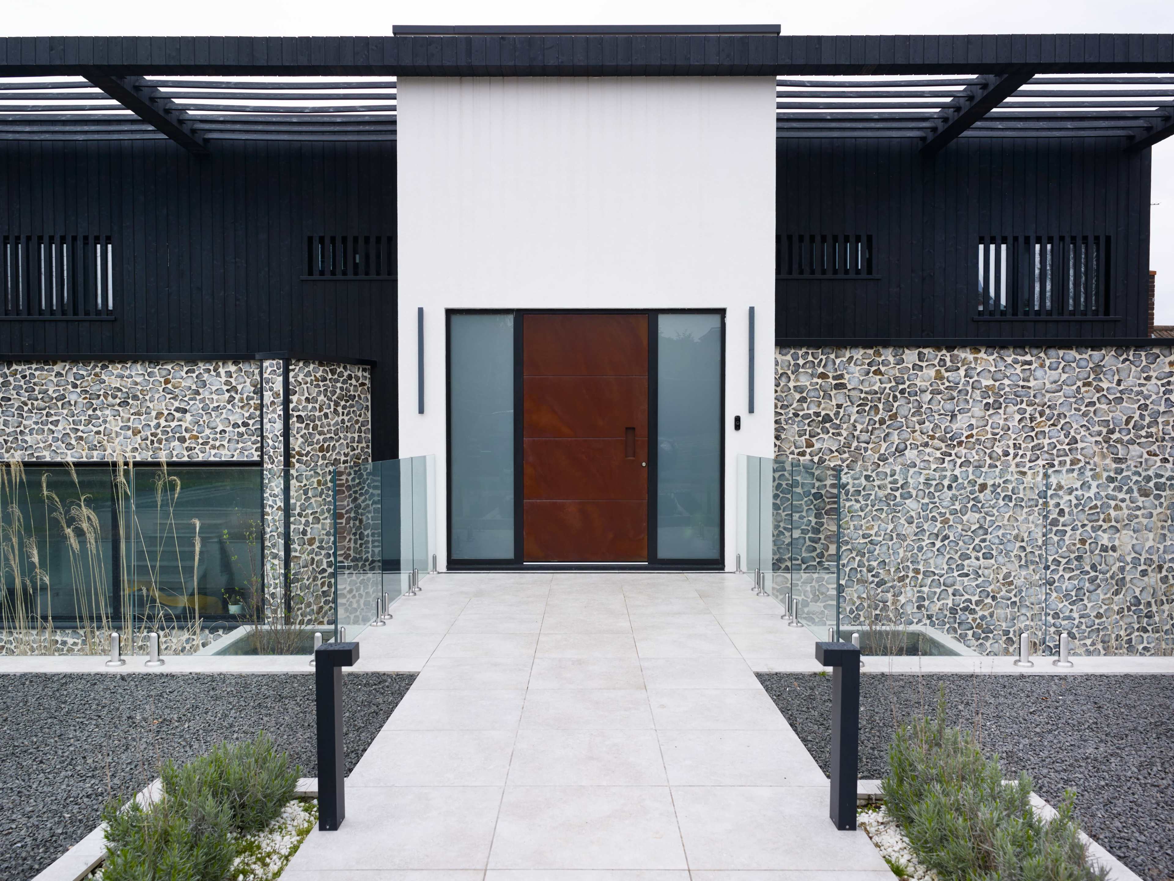 A modern house with a dark exterior, featuring a large wooden door, stone walls, and a pathway lined with glass railings and gravel.
