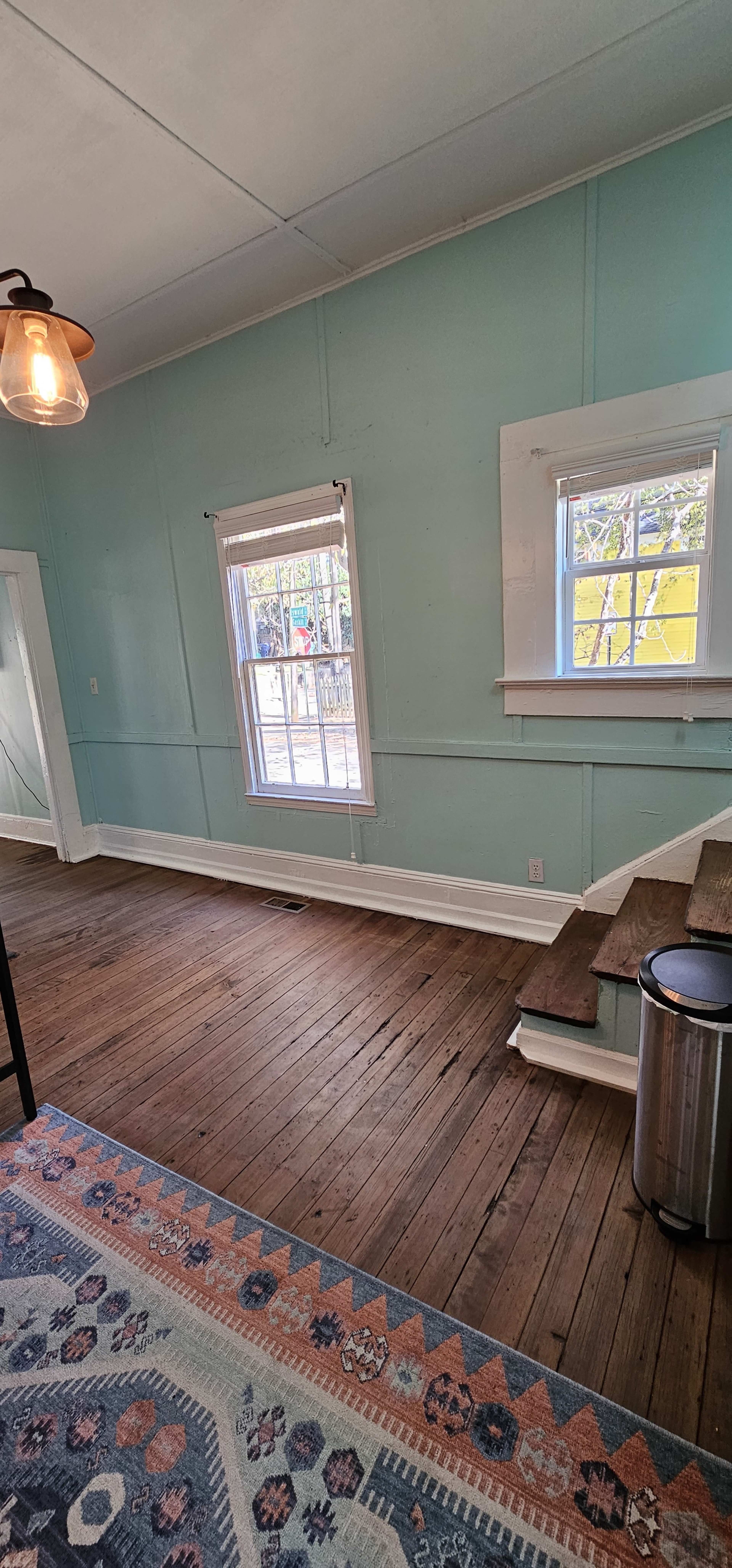 A room with light blue walls, two windows, a wooden floor, and a staircase leading to a lower area.