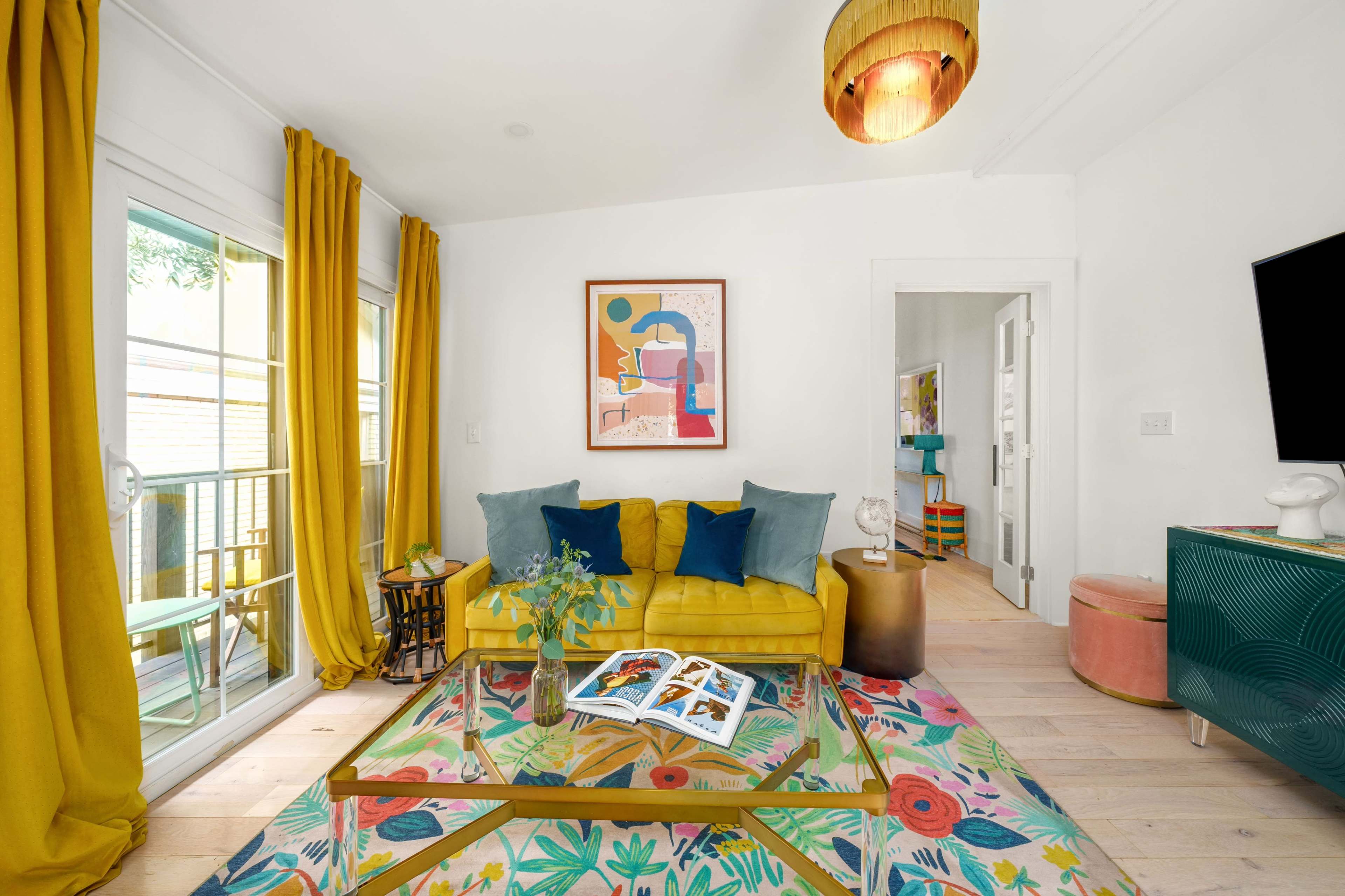 A bright living room features a yellow sofa with pillows, a glass coffee table, and large windows adorned with yellow curtains.