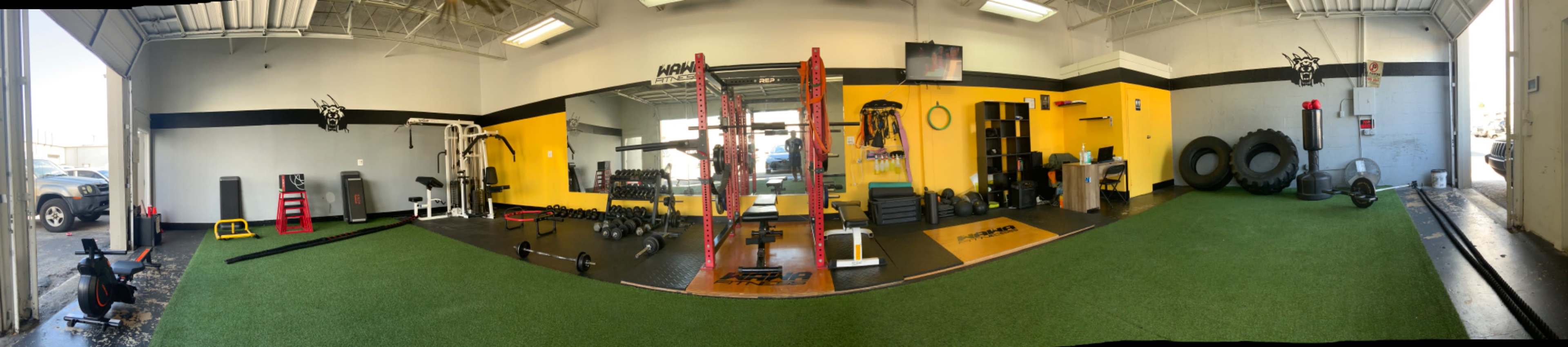 The image shows a wide view of a gym with various exercise equipment, including weightlifting stations, a rowing machine, and free weights, set against walls painted in black and yellow.