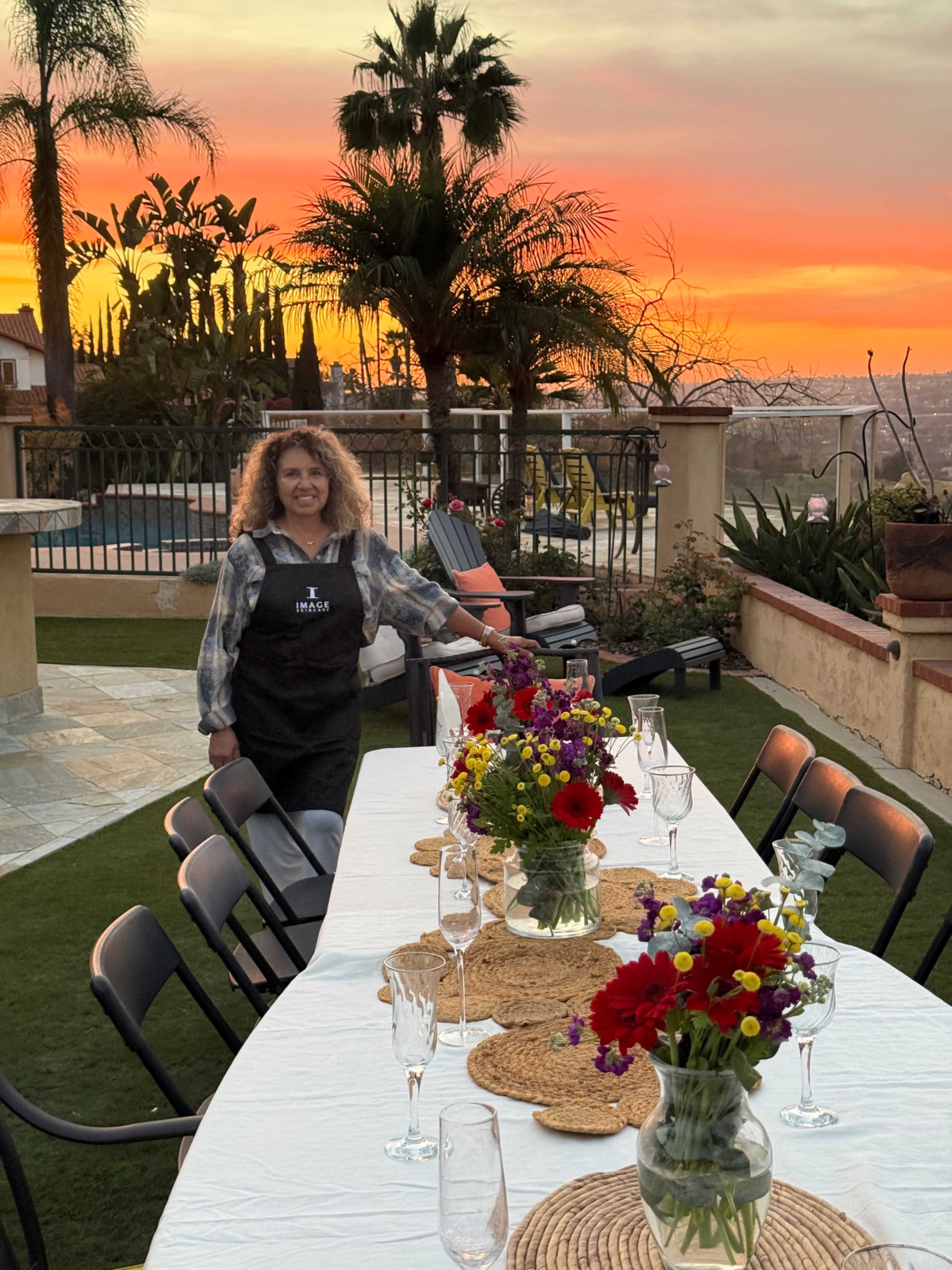 A woman wearing an apron stands next to a long table set with colorful flower arrangements under a vibrant sunset.