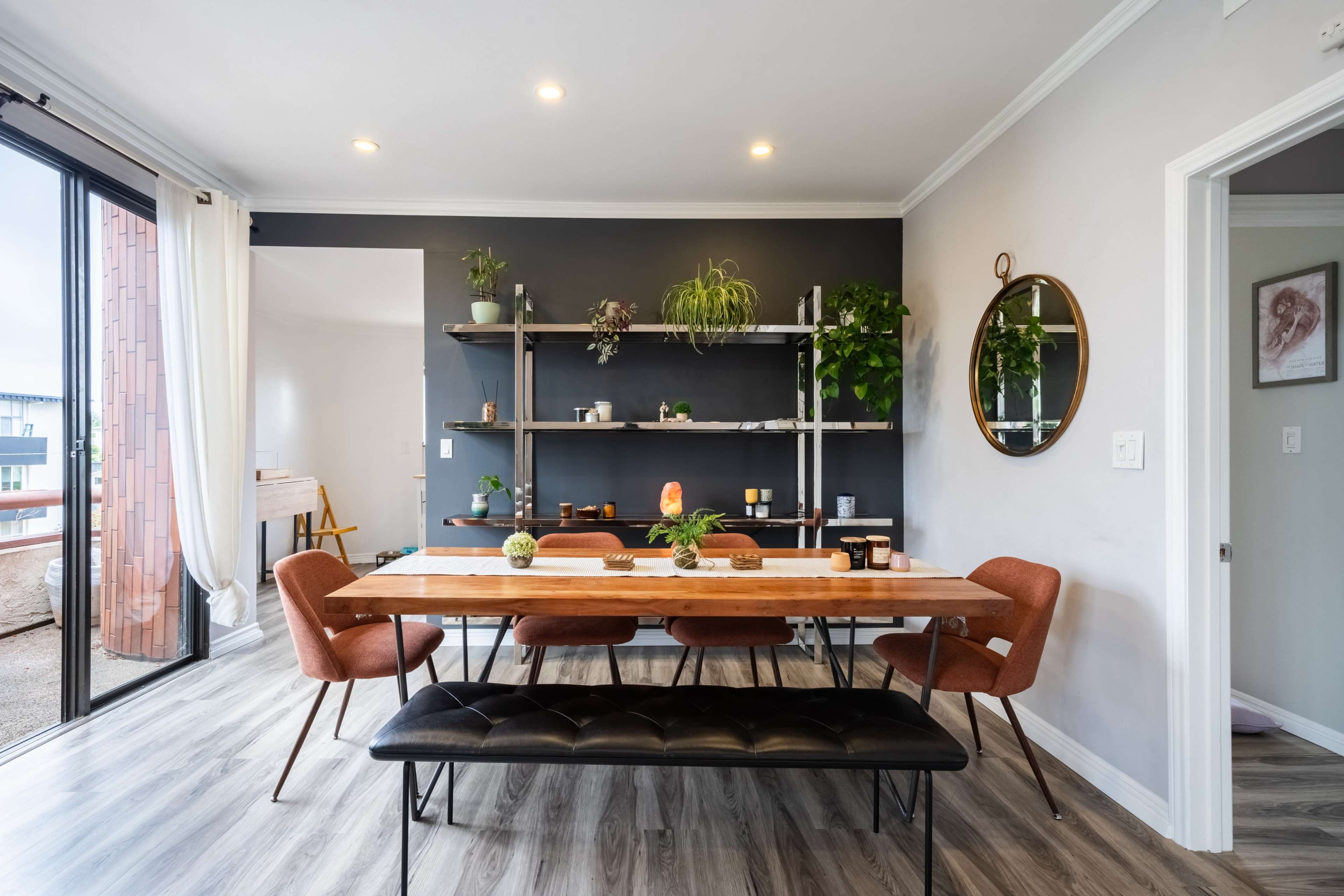 A modern dining room with a long wooden table, surrounded by orange chairs, and adorned with plants and decorative items on shelves.