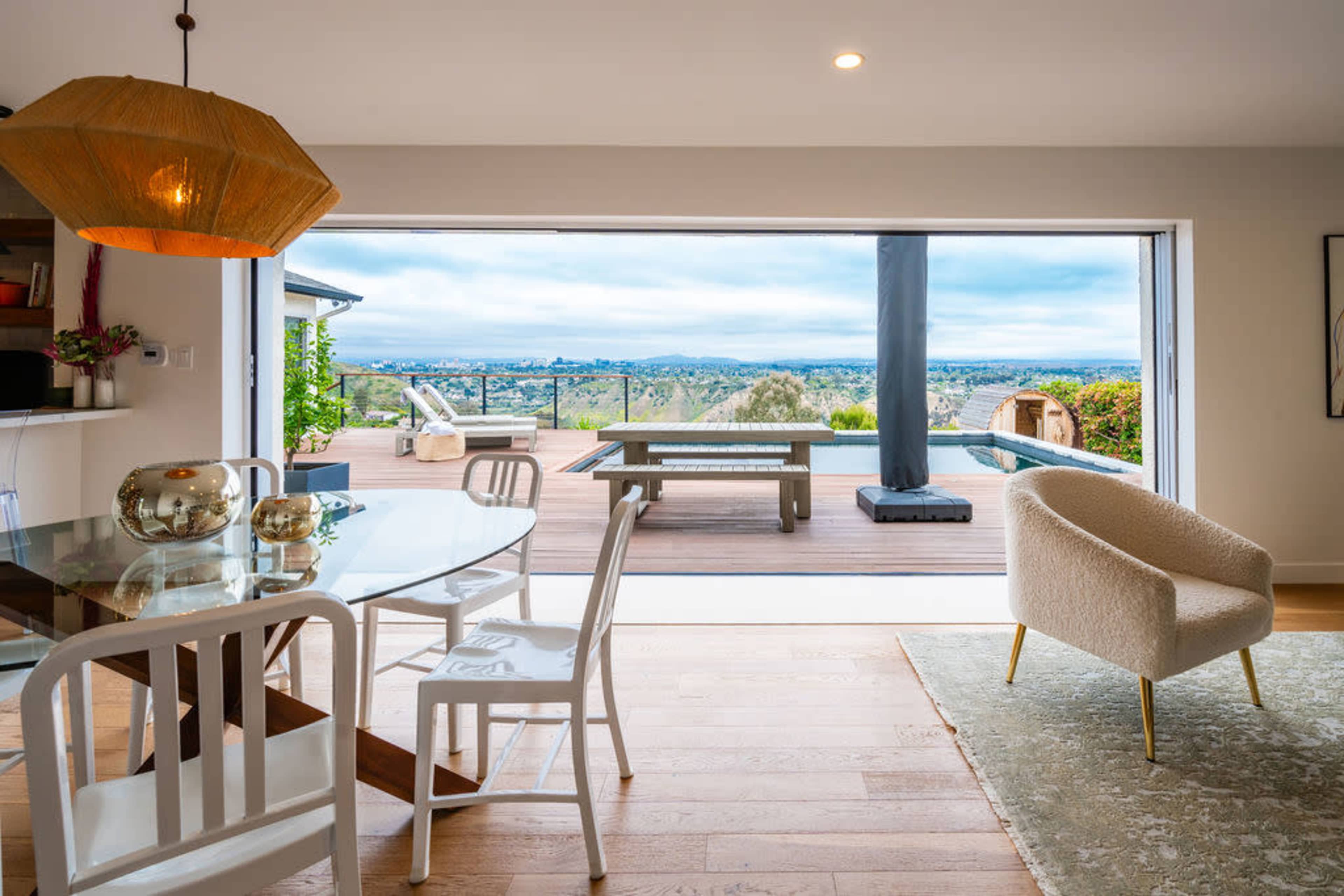 A dining area features a glass table and white chairs, with a large sliding door that opens to a wooden deck overlooking a panoramic view.