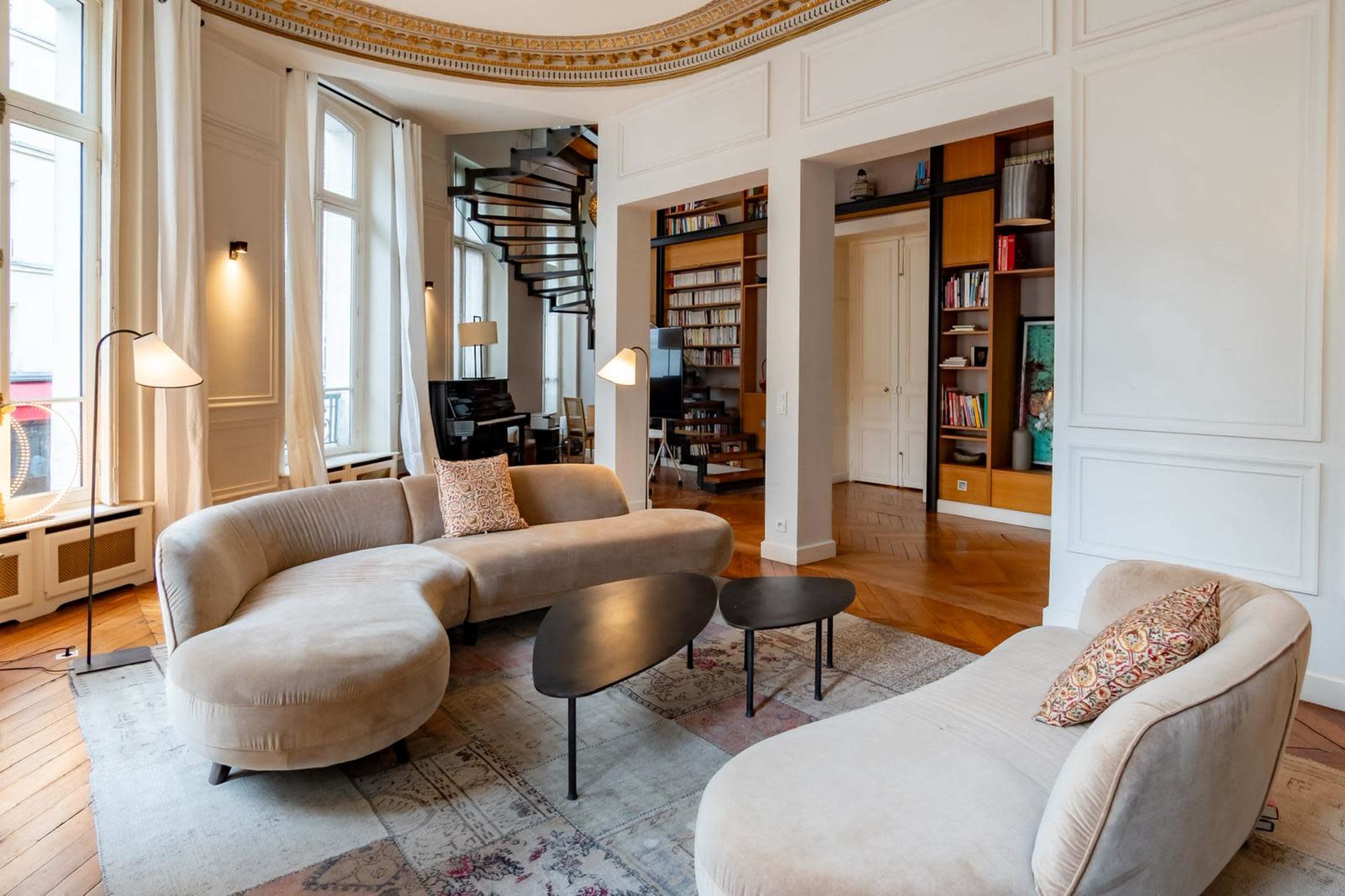The image shows a modern living room with curved sofas, a black coffee table, and a spiral staircase leading to an upper level, all framed by large windows.