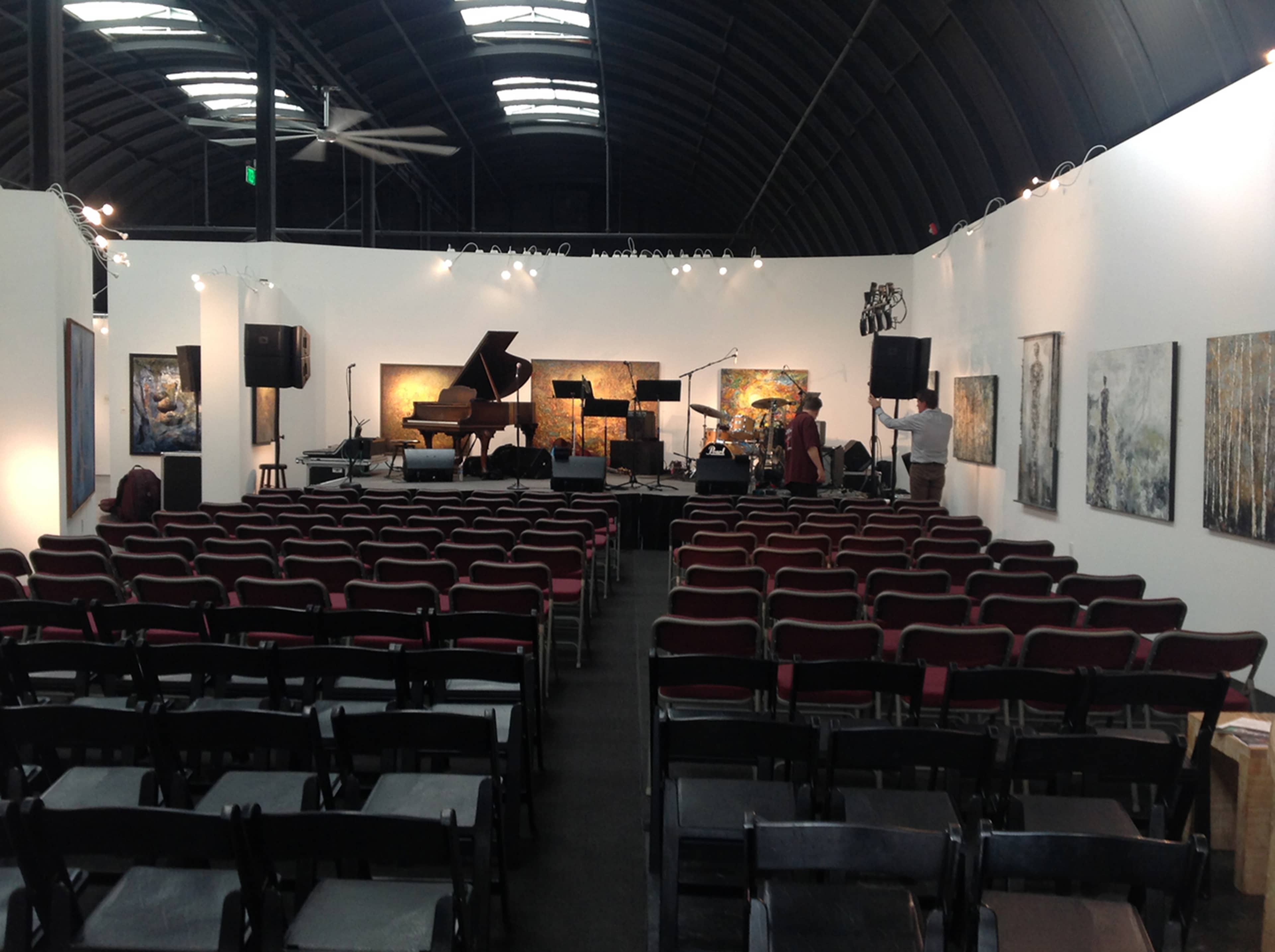 An empty performance space with rows of black chairs facing a grand piano and a backdrop of artwork on the walls.