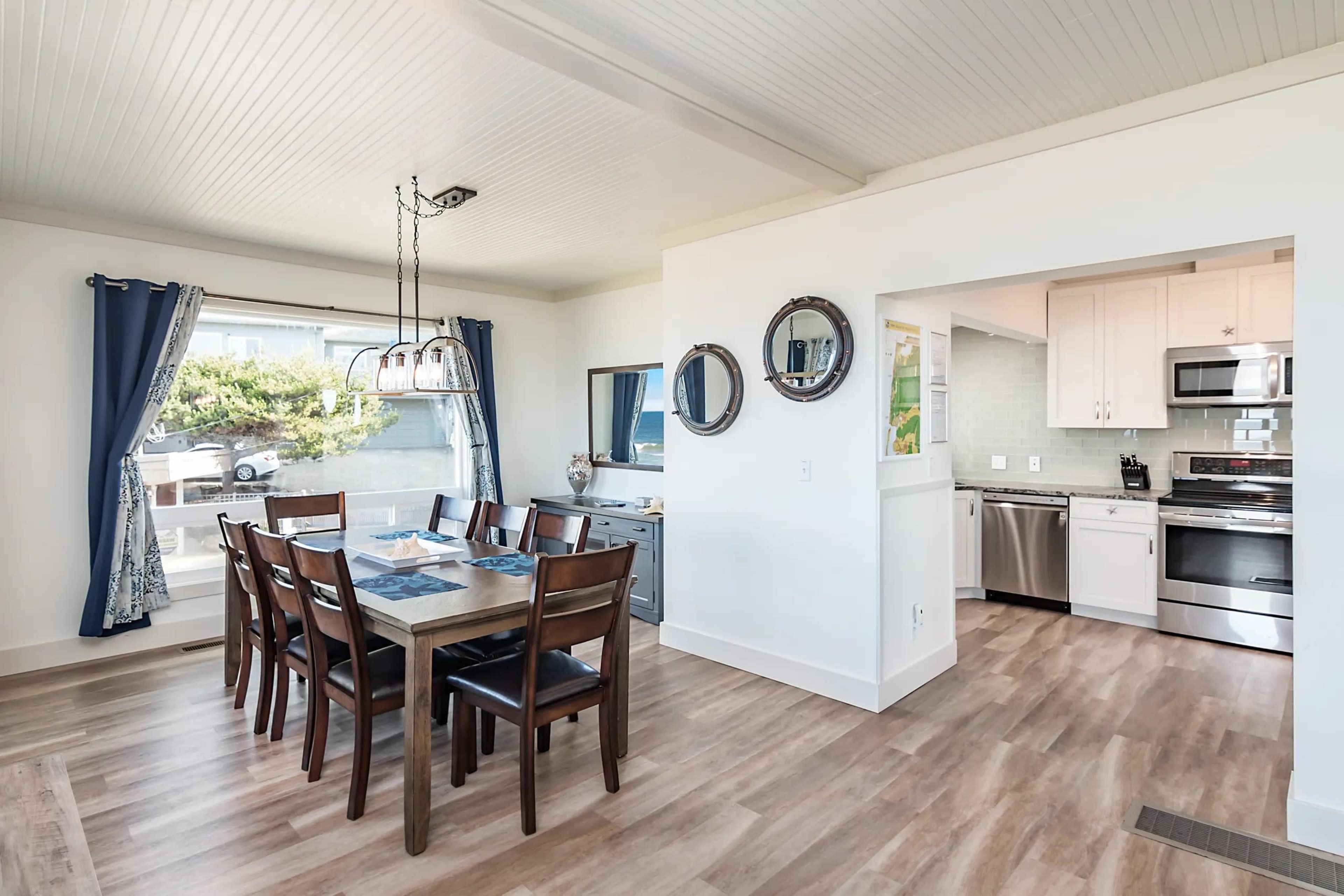 The image shows a dining area with a long wooden table surrounded by chairs, adjacent to a kitchen with modern appliances and a large window providing natural light.