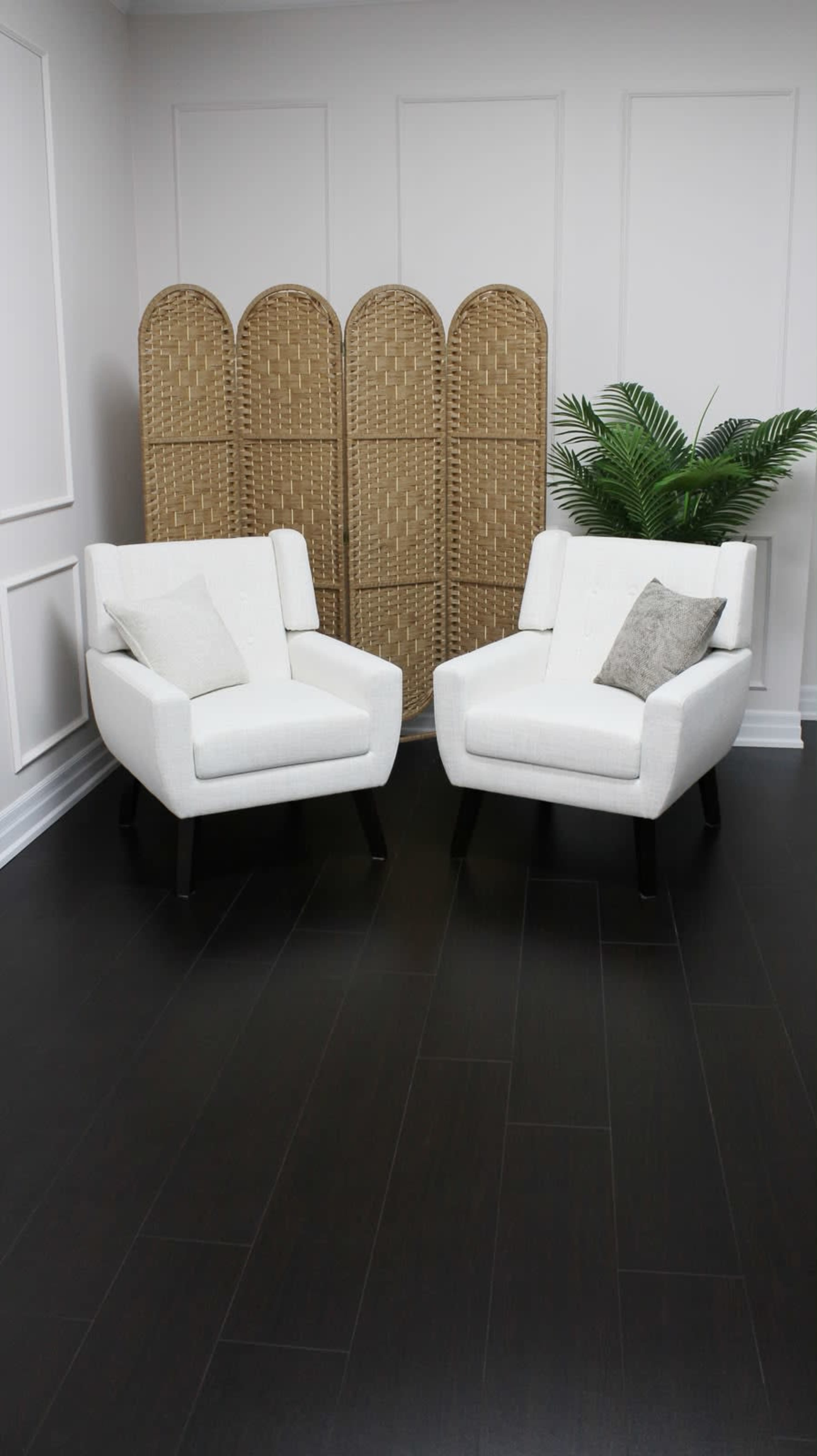 The image shows two modern white armchairs with dark wooden legs positioned facing each other in a room with a decorative screen and a potted plant in the background.