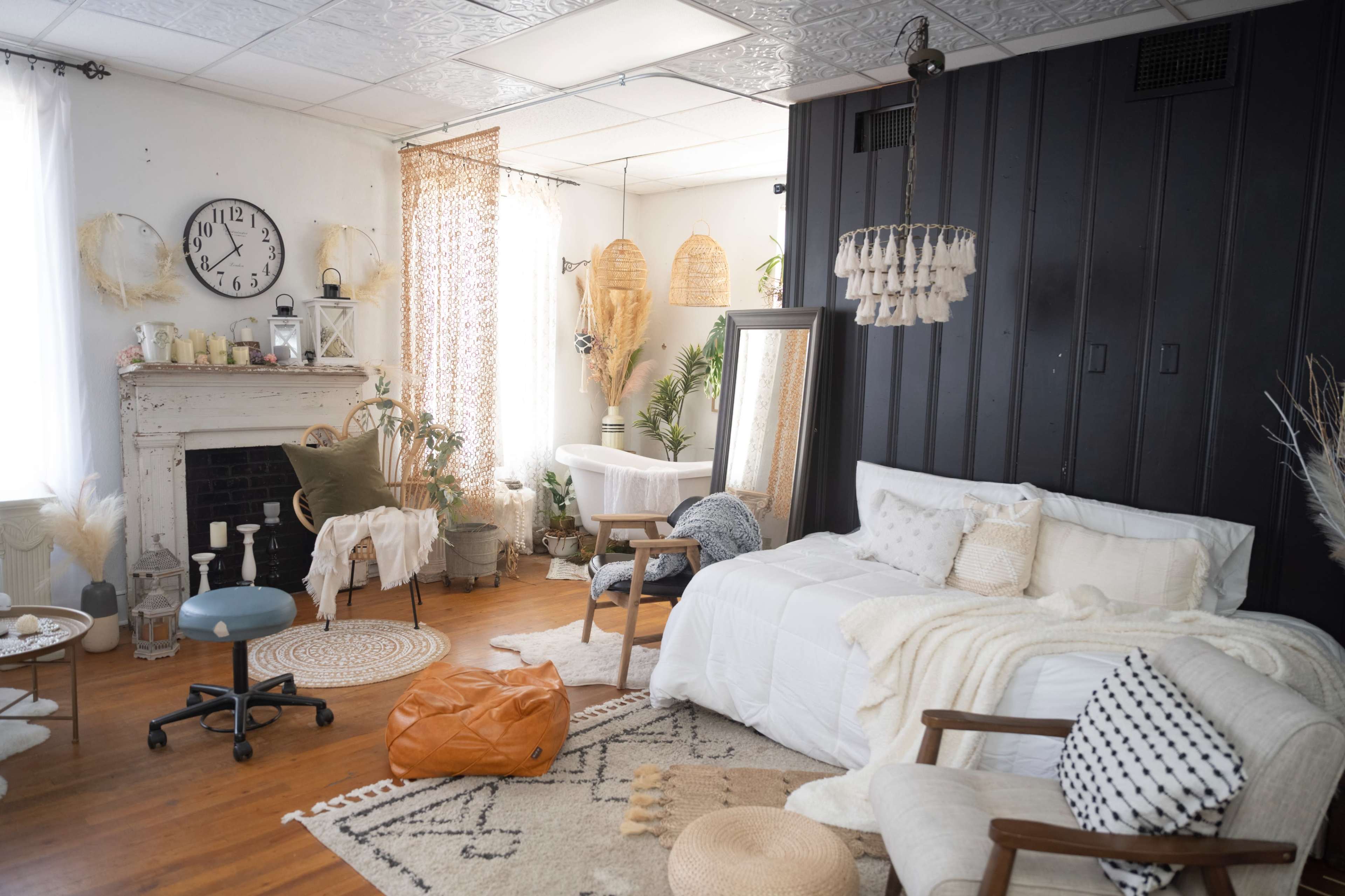 The image shows a cozy room featuring a bed, armchairs, decorative plants, a clock on the wall, and various textiles arranged aesthetically.