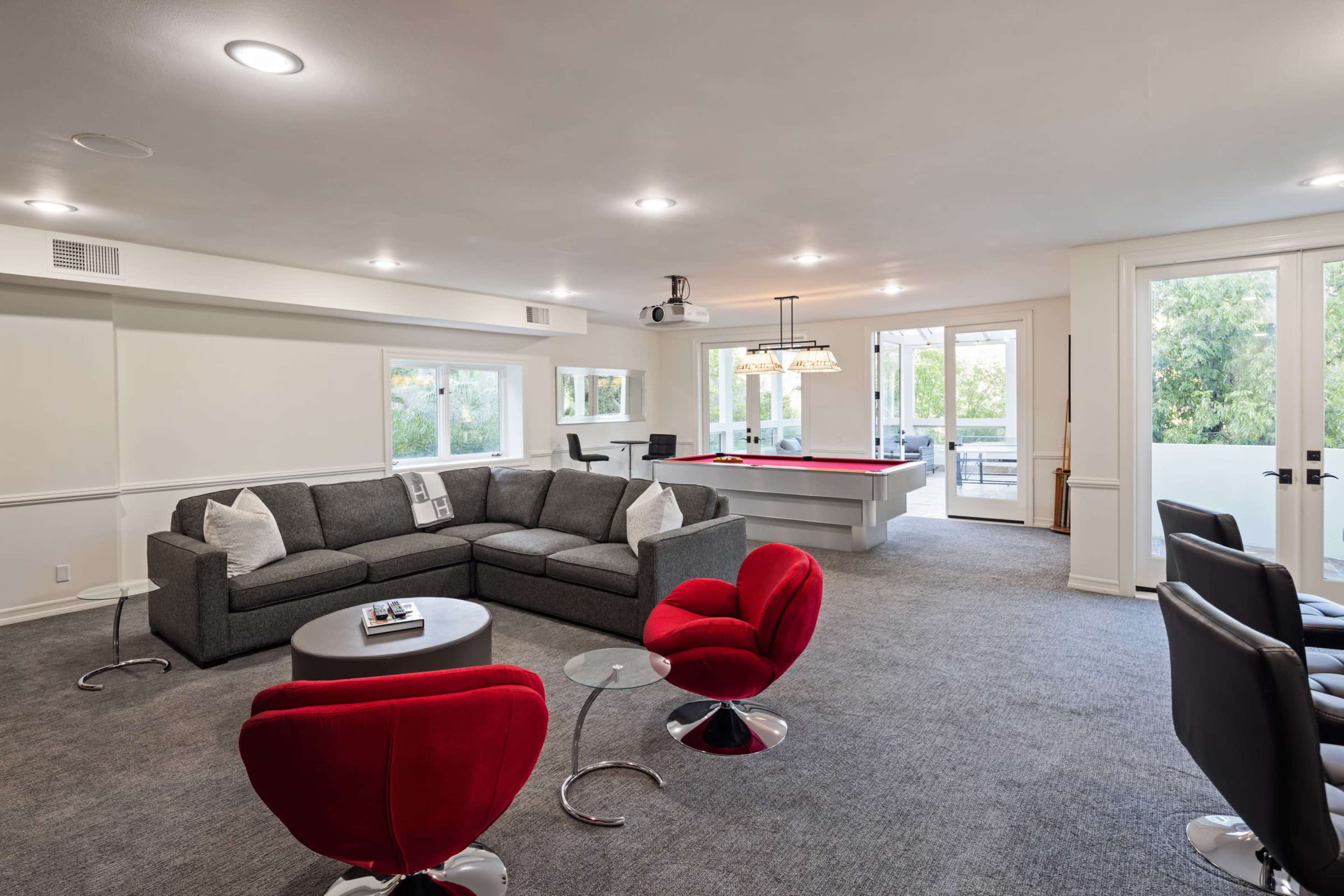 A spacious living area features a gray sectional sofa, red accent chairs, a round coffee table, and large windows that open to an outdoor space.