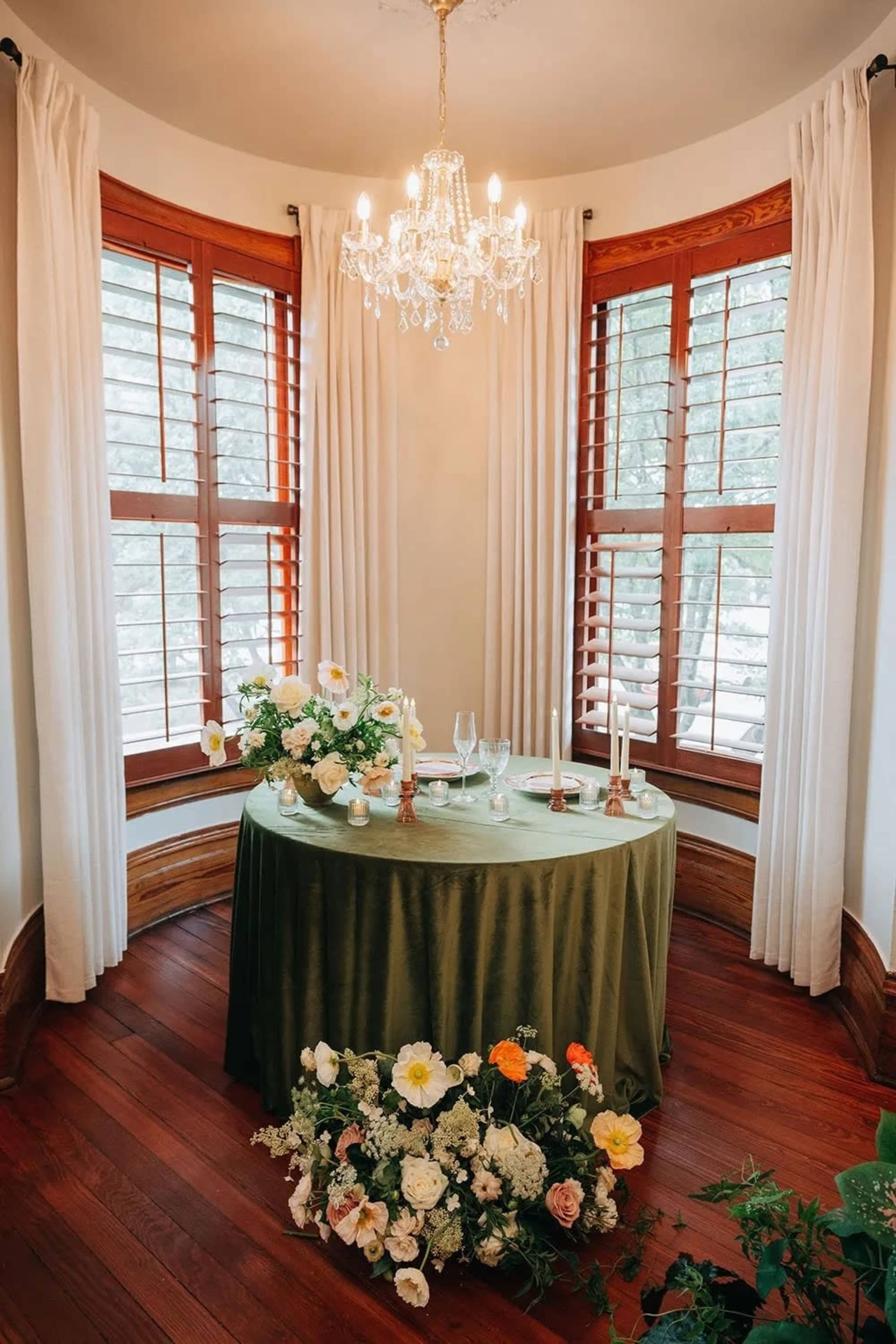 A round, green table is adorned with flowers and candles in a well-lit corner of a room featuring large windows with white curtains.