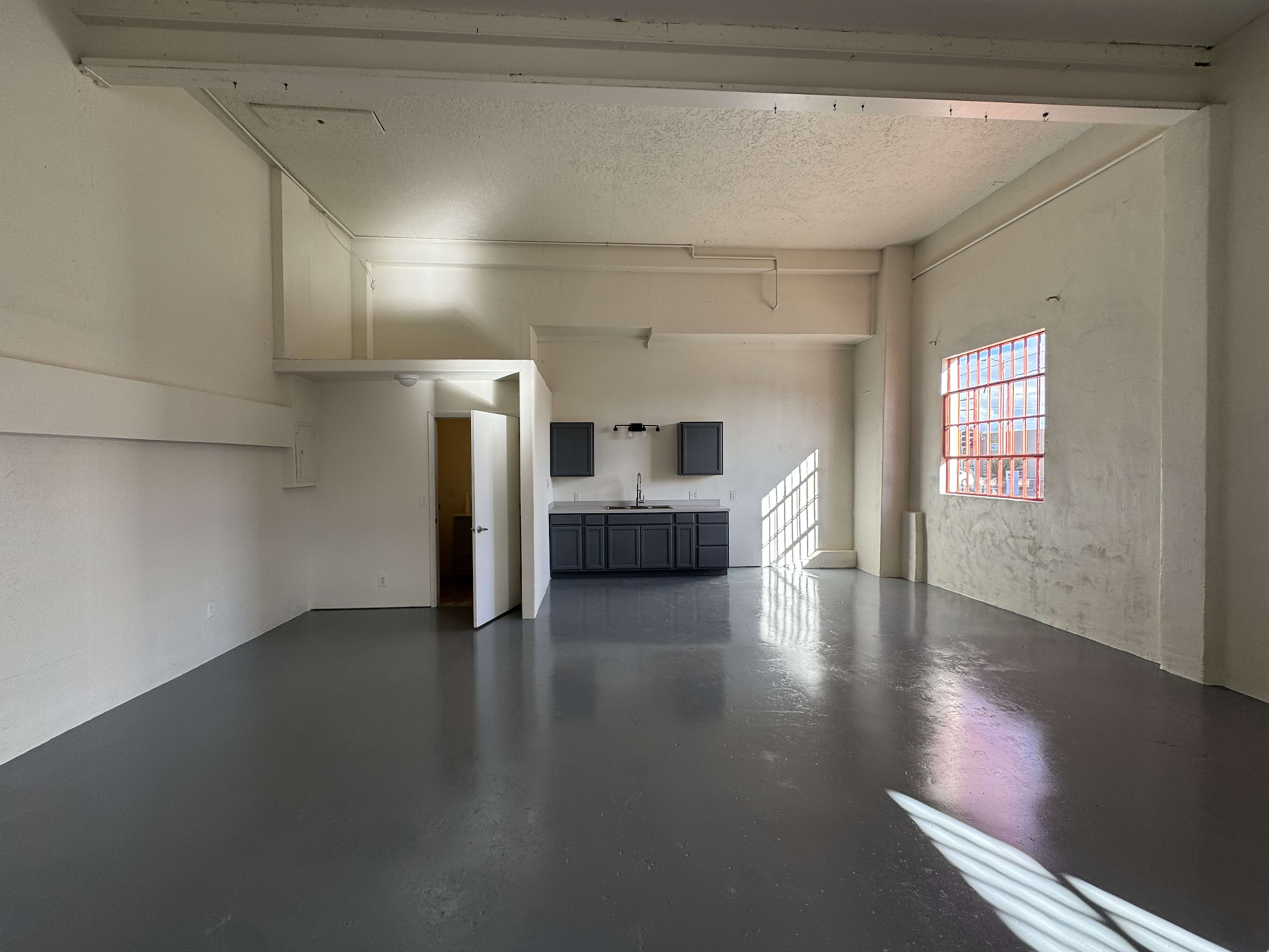 A spacious, vacant room with polished gray floors, white walls, and a kitchenette area featuring dark cabinets and two windows allowing natural light to enter.