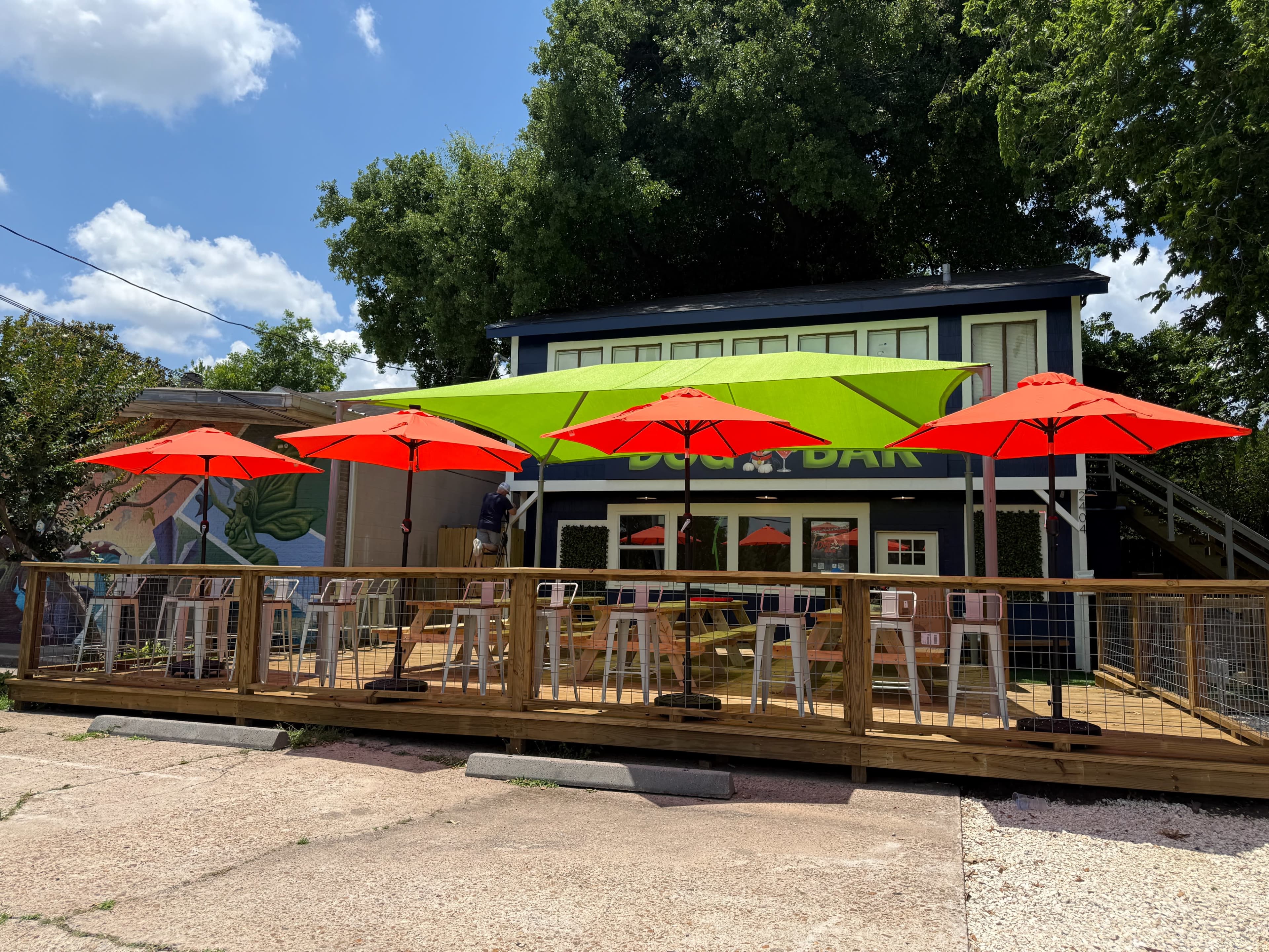 The image shows a wooden deck with bright red umbrellas overhead, leading up to a two-story building labeled as a bar, surrounded by trees and blue sky.