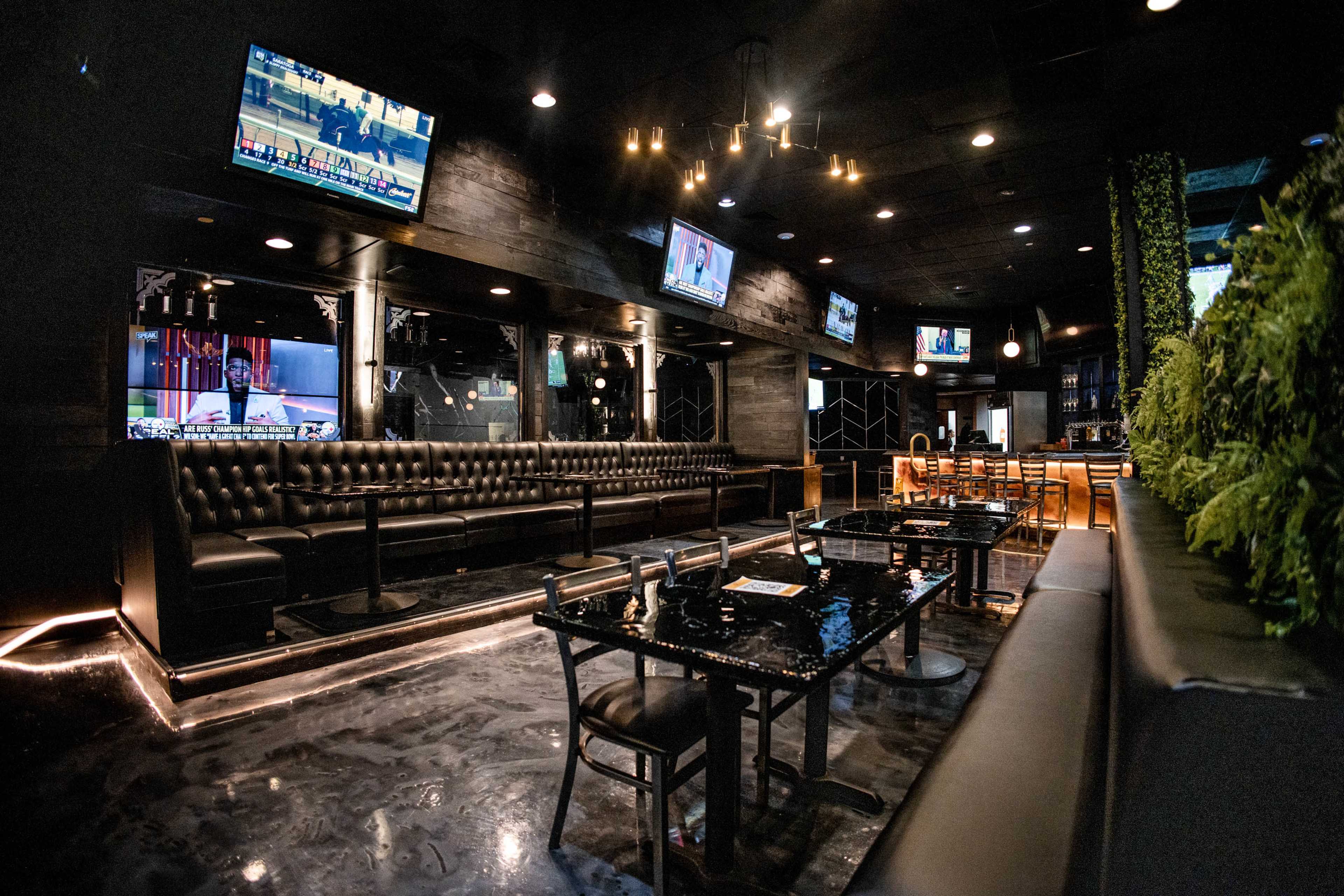 The image shows a modern, dimly lit interior of a bar or restaurant with black leather seating, several televisions mounted on the walls, and tables arranged throughout the space.