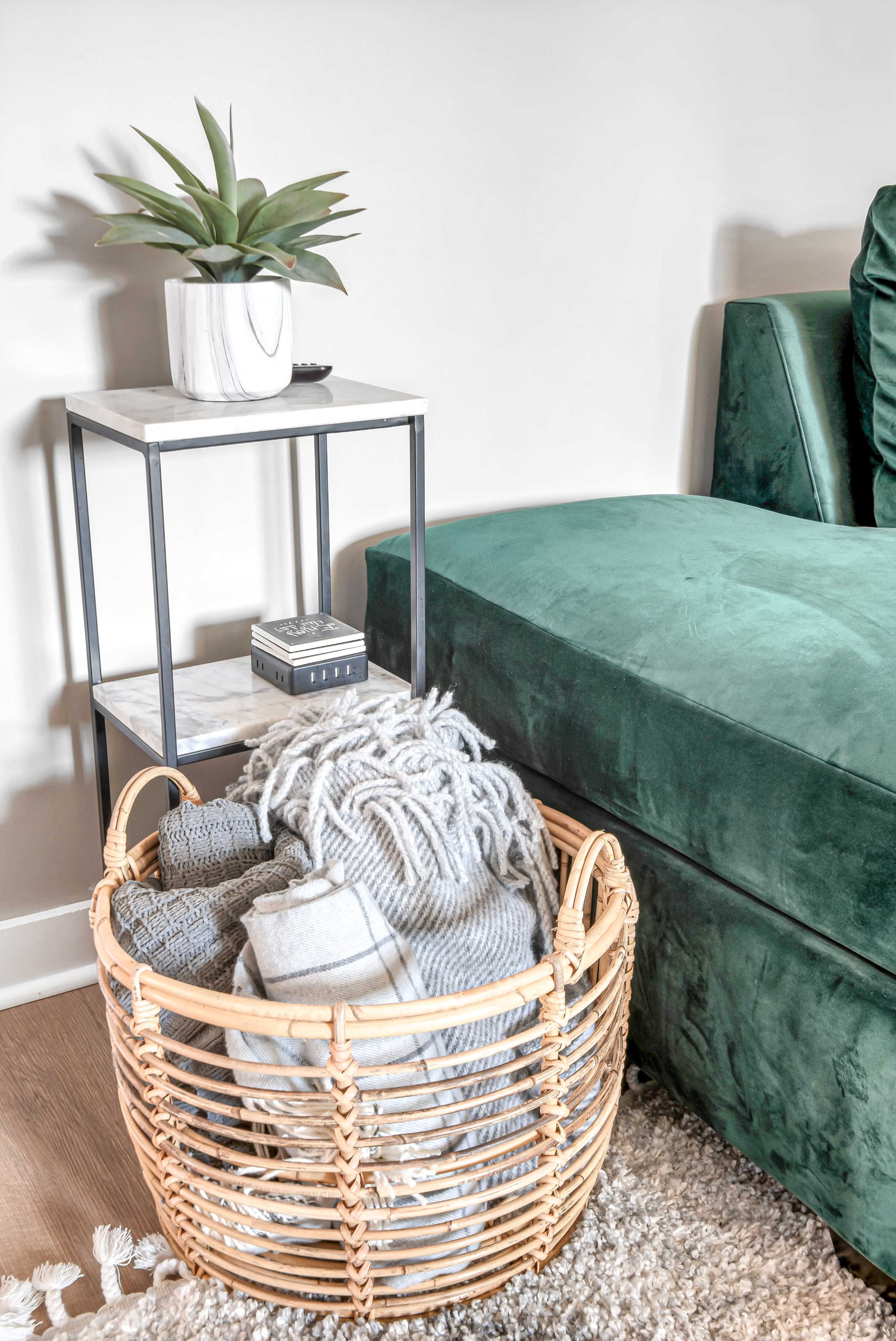 A wicker basket filled with folded blankets sits beside a small table featuring a potted plant and a decorative box, next to a green upholstered sofa.