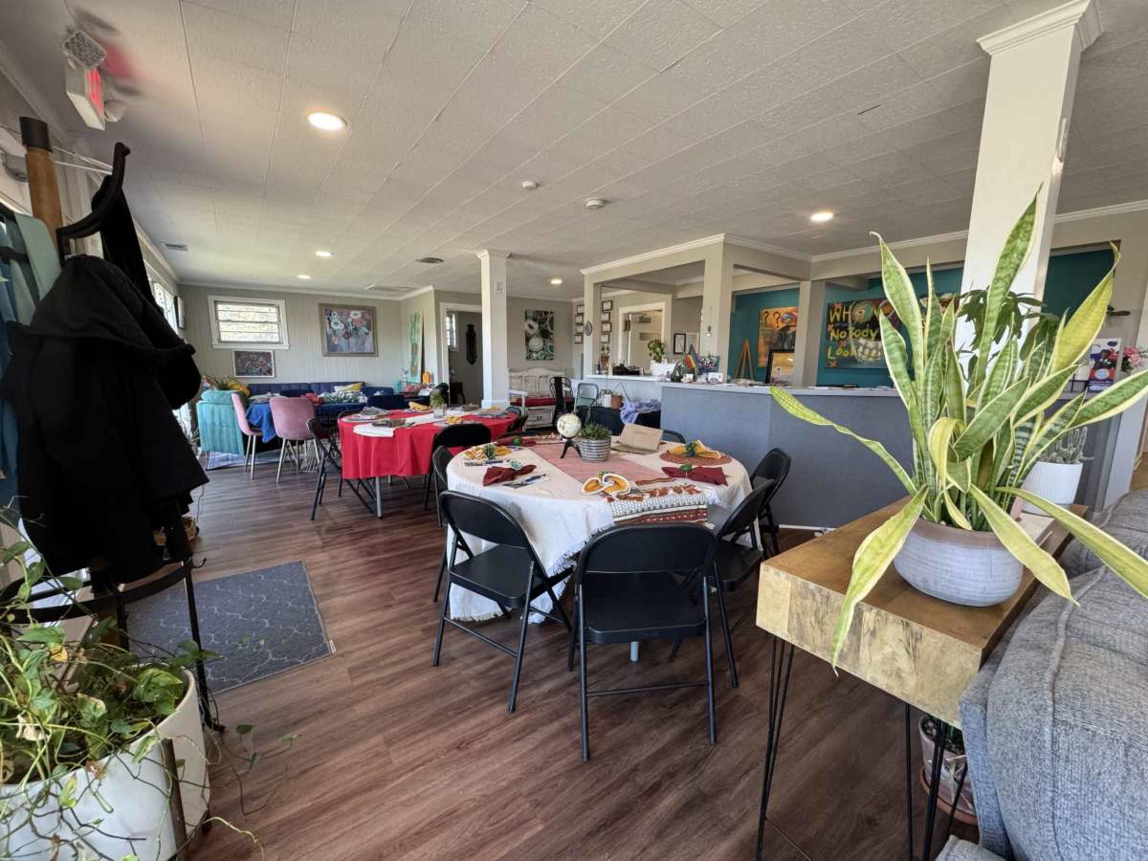The image shows a spacious dining area with several tables set for guests, each decorated differently, along with plants and various furnishings in a well-lit environment.