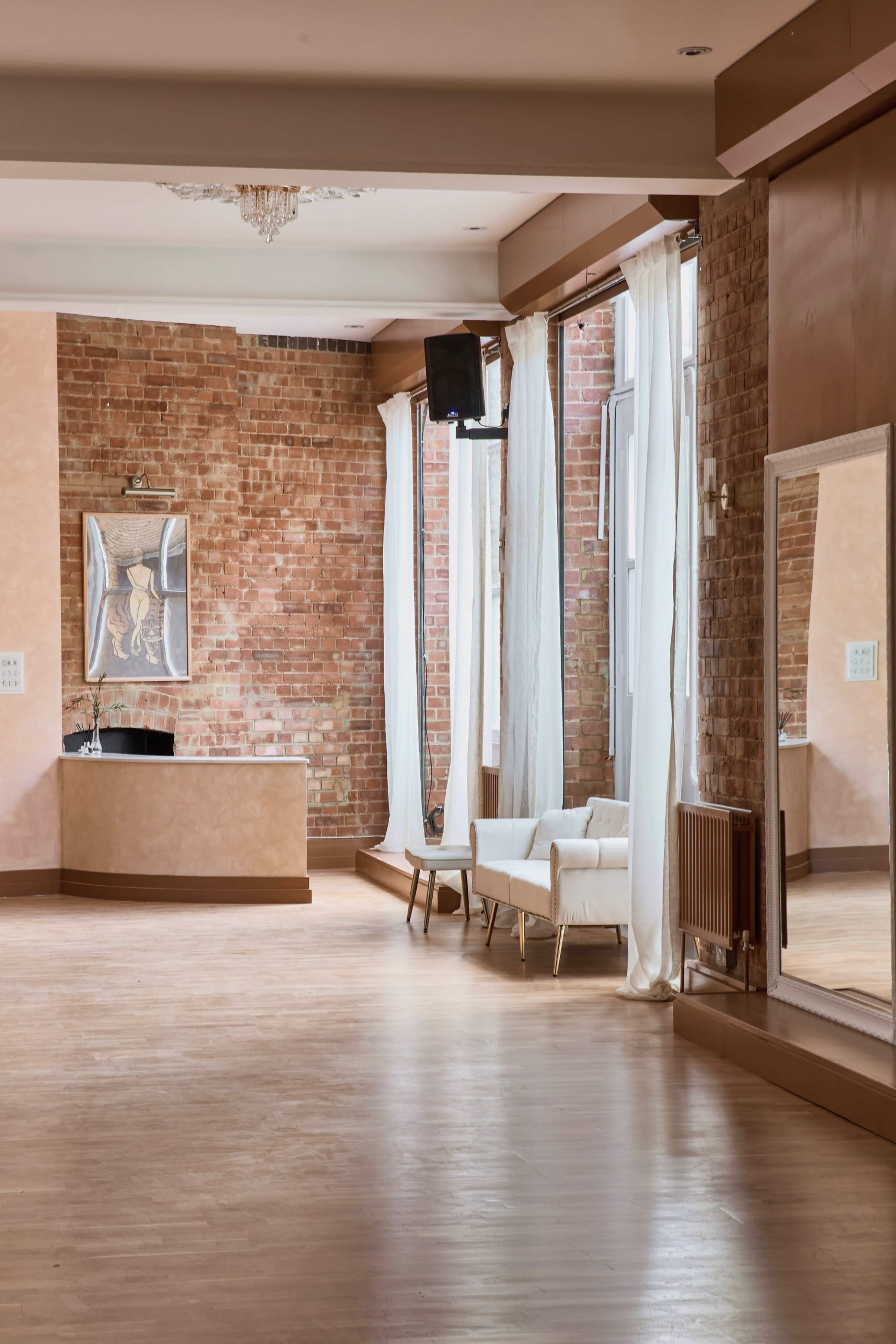 The interior of a brightly lit dance studio features exposed brick walls, large windows with sheer curtains, and a white couch beside a tall mirror.