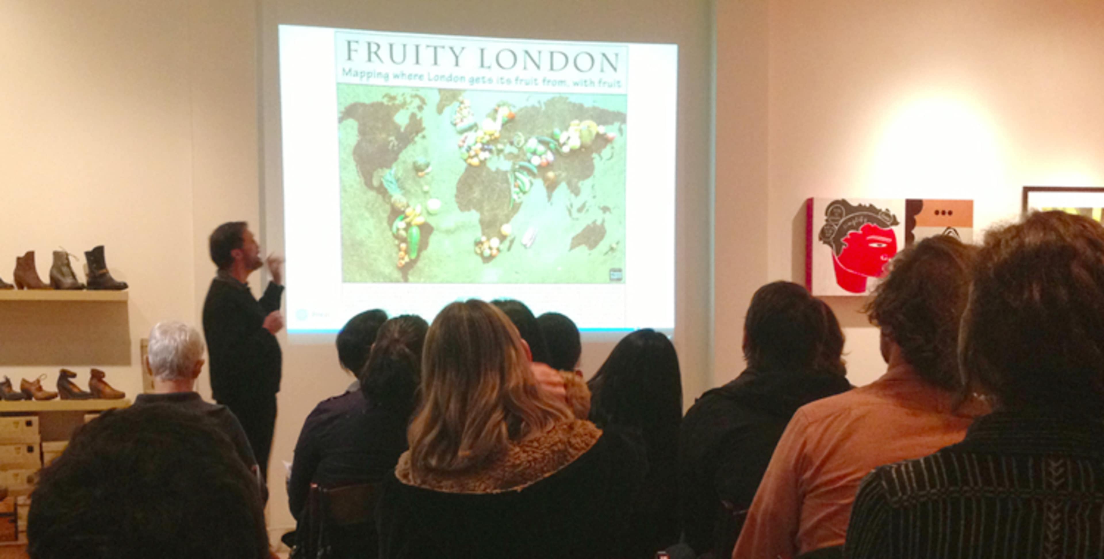 A speaker stands in front of a projection titled "Fruity London" while an audience observes attentively in a gallery setting.