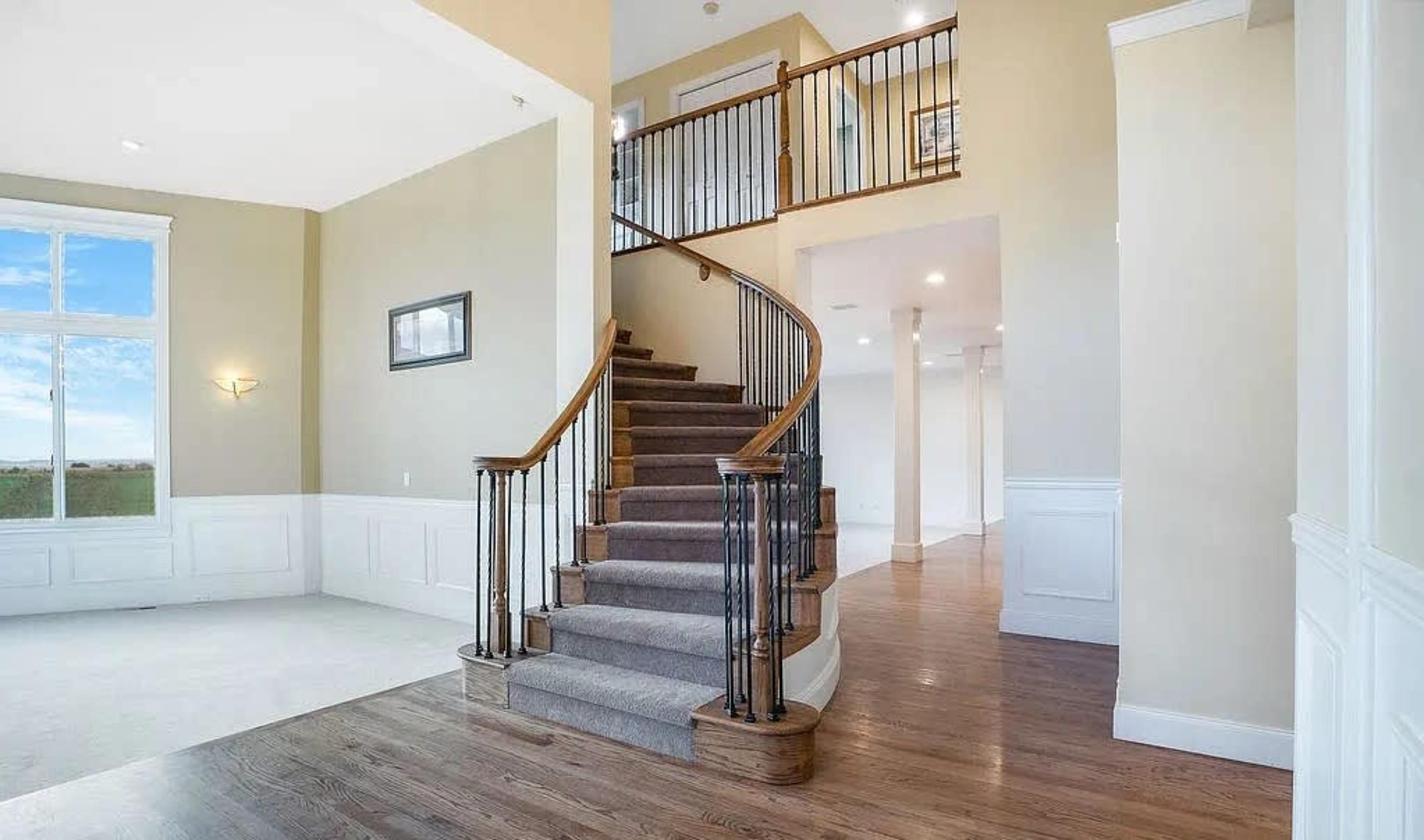 A curved staircase with a wooden handrail sits in a spacious entryway featuring light-colored walls and large windows.