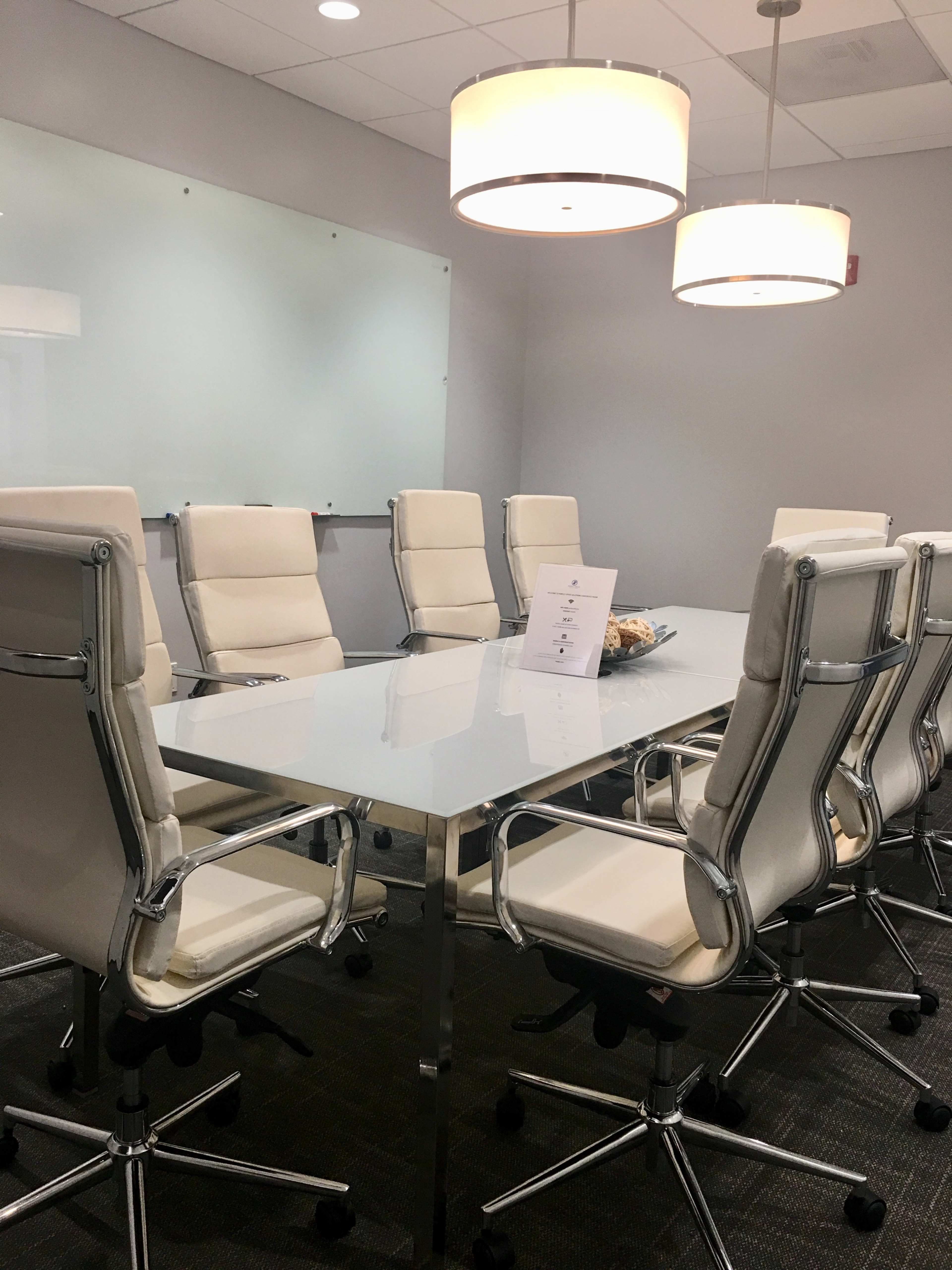 A modern conference room features a glass-topped table surrounded by white leather chairs and illuminated by pendant lights.