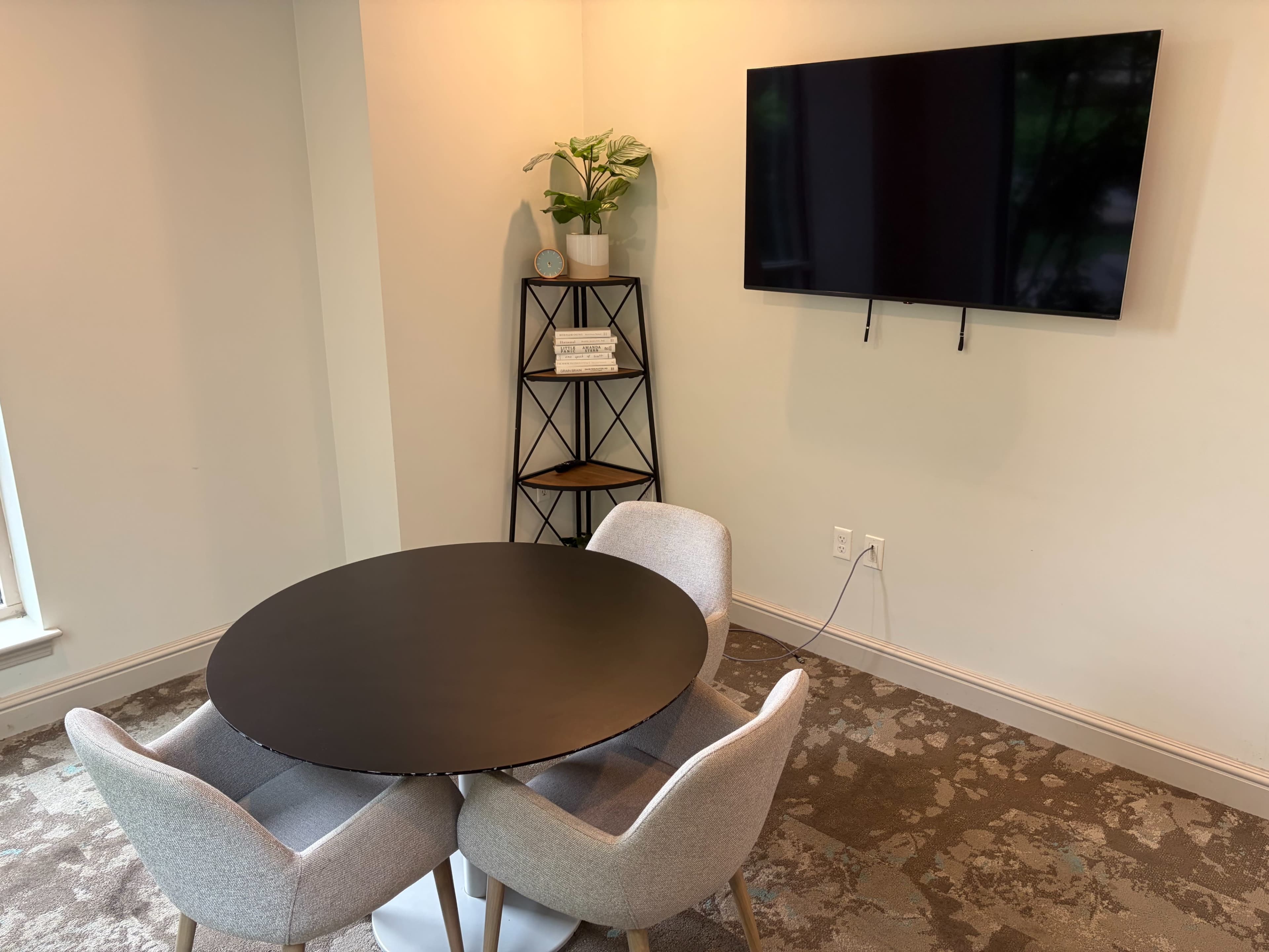 The image shows a small dining area featuring a round black table with two gray chairs, a wall-mounted television, and a minimalist shelf with decorative items and a plant.