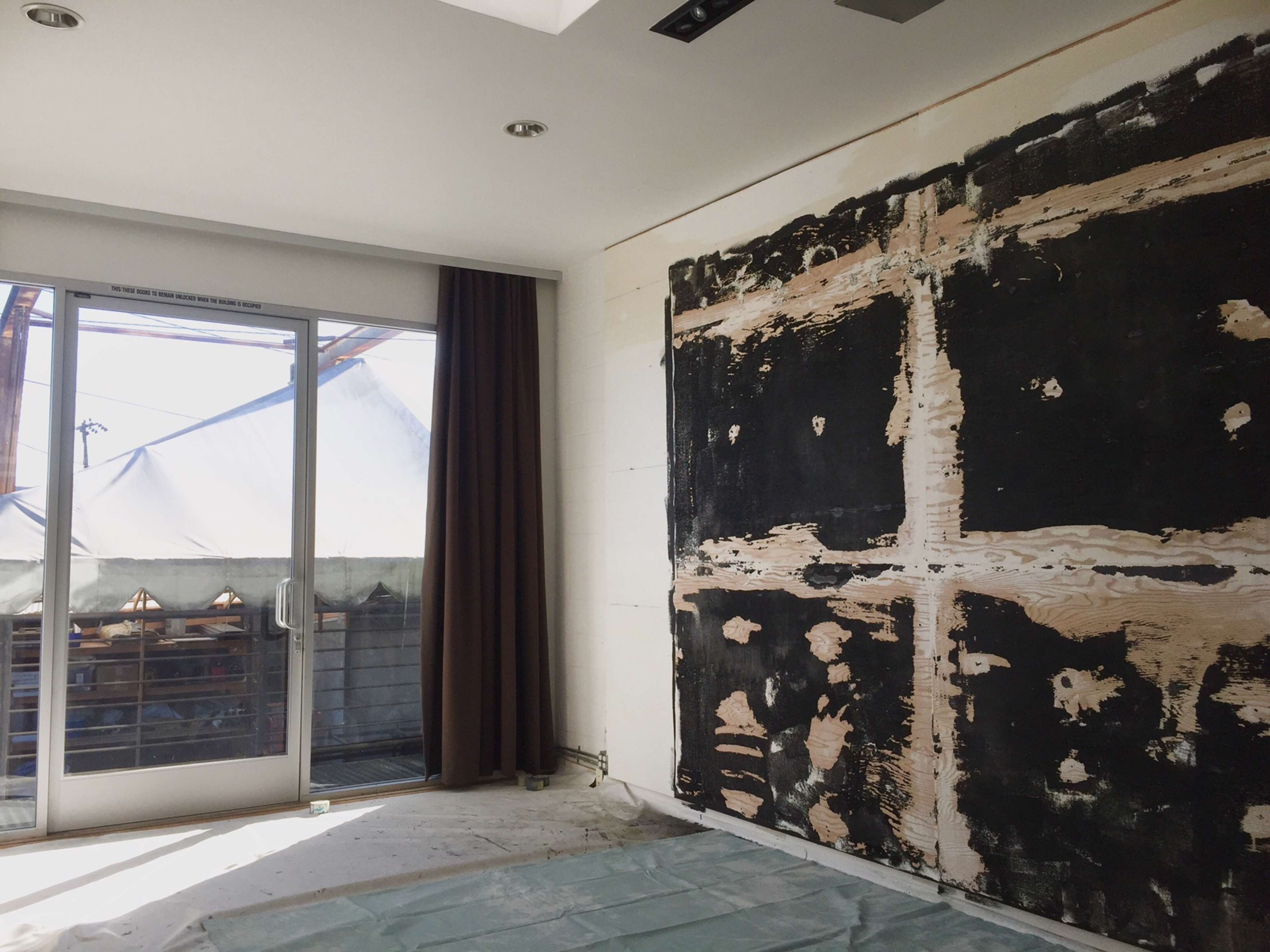 The image shows an unfinished interior room with bare wall patches and a large window revealing a tented outdoor space.