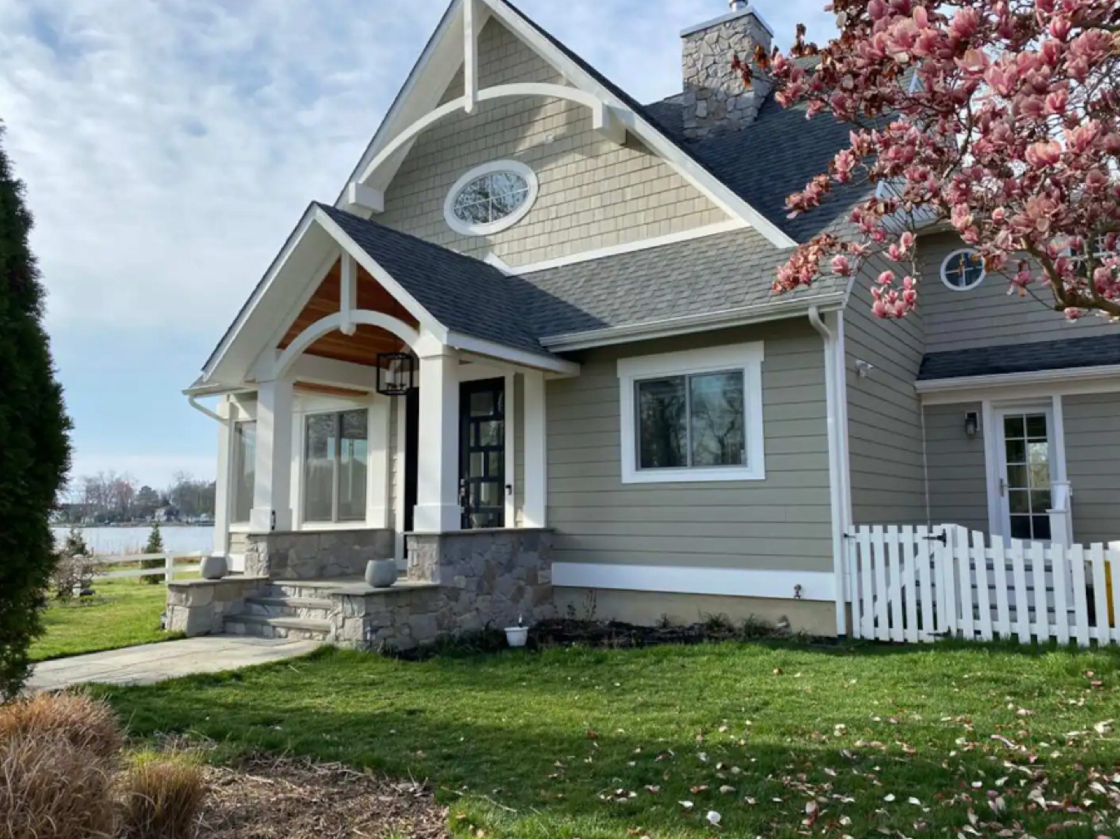 A two-story house with a light gray exterior, a front porch supported by pillars, and a white picket fence surrounded by landscaped green grass and a blooming tree in the foreground.