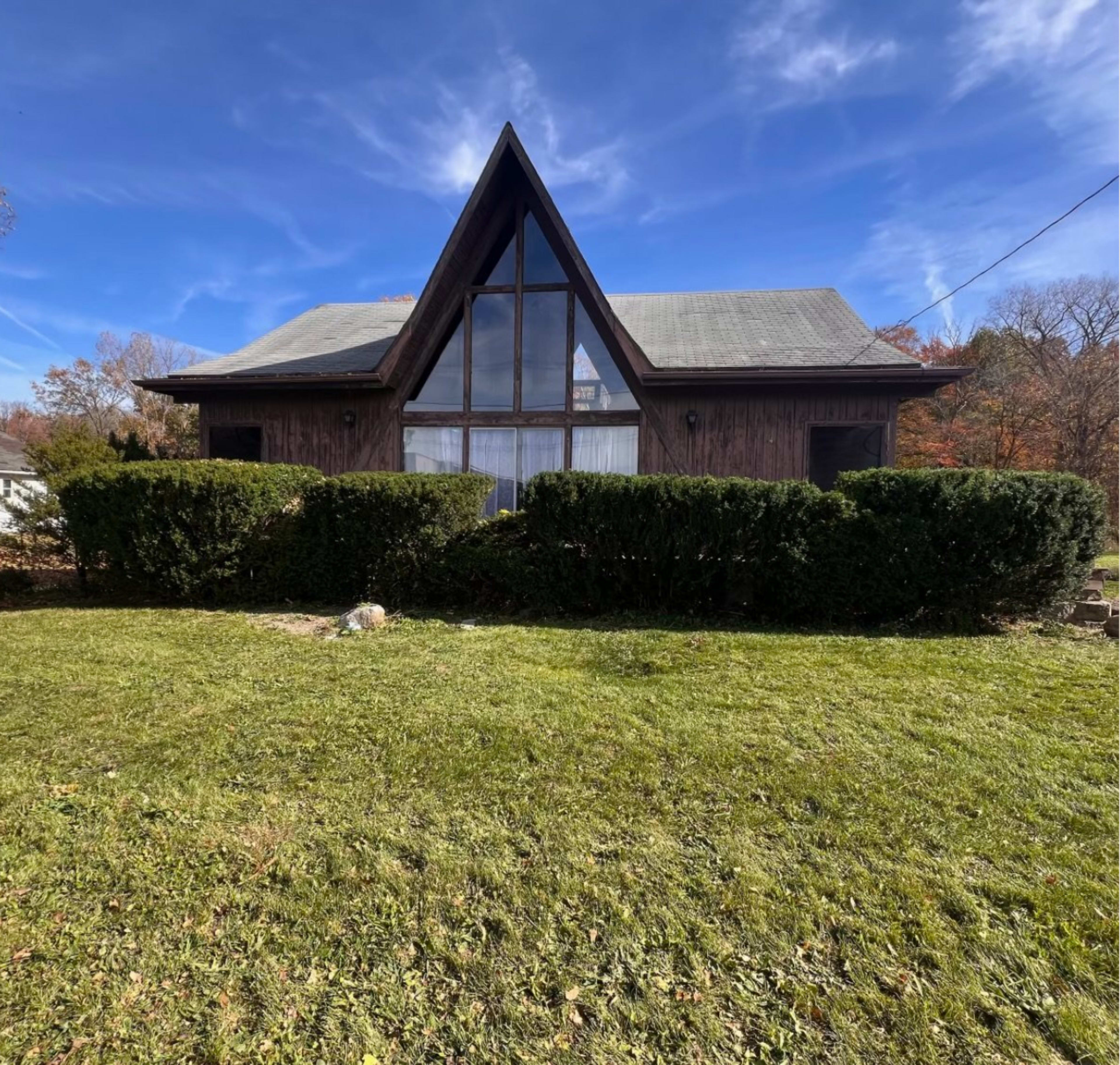 A brown wooden house with a steeply angled roof and large front windows is surrounded by neatly trimmed hedges and a grassy lawn.