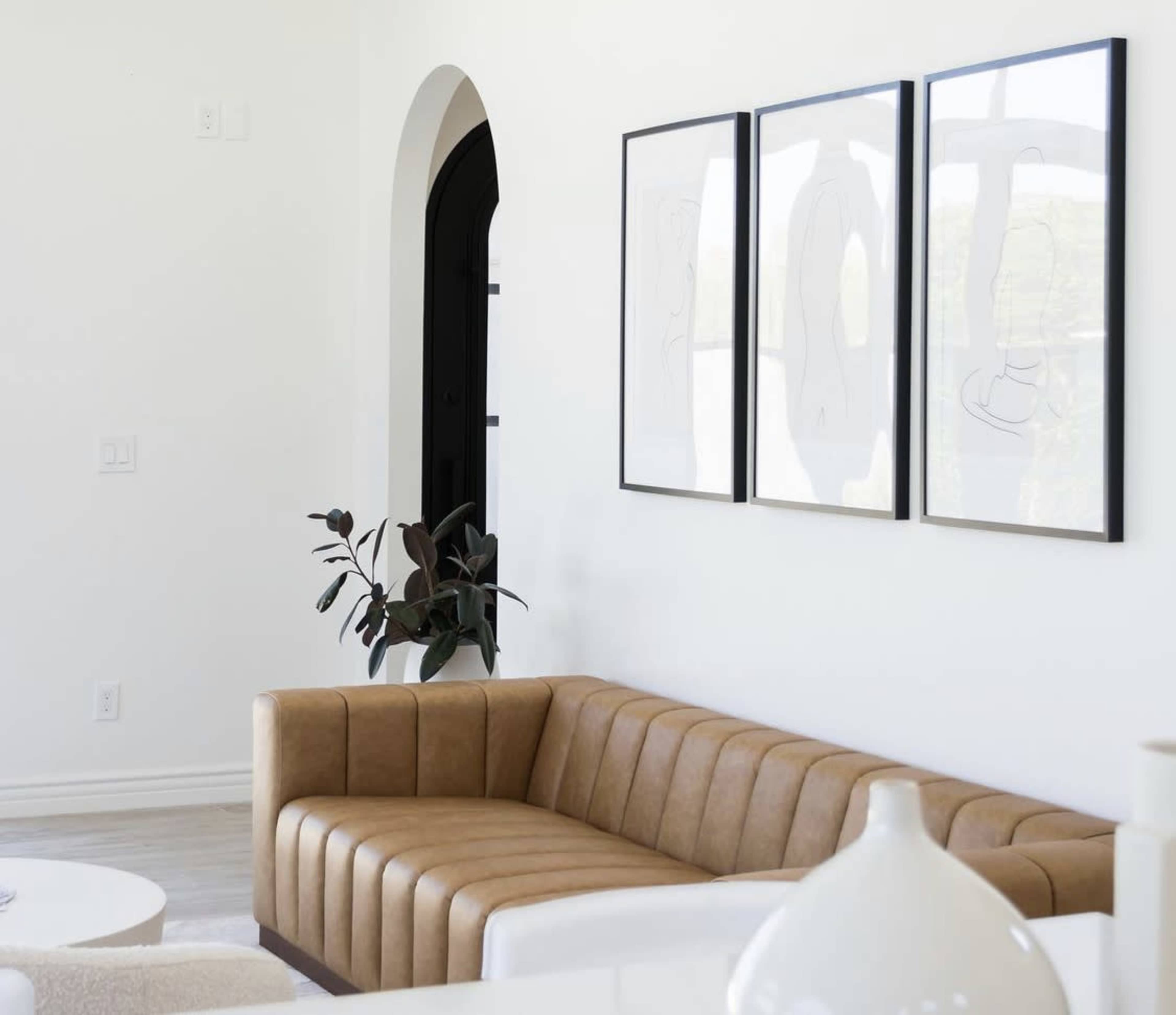 A tan leather sofa is positioned against a white wall, complemented by three framed minimalist artworks and a potted plant nearby.