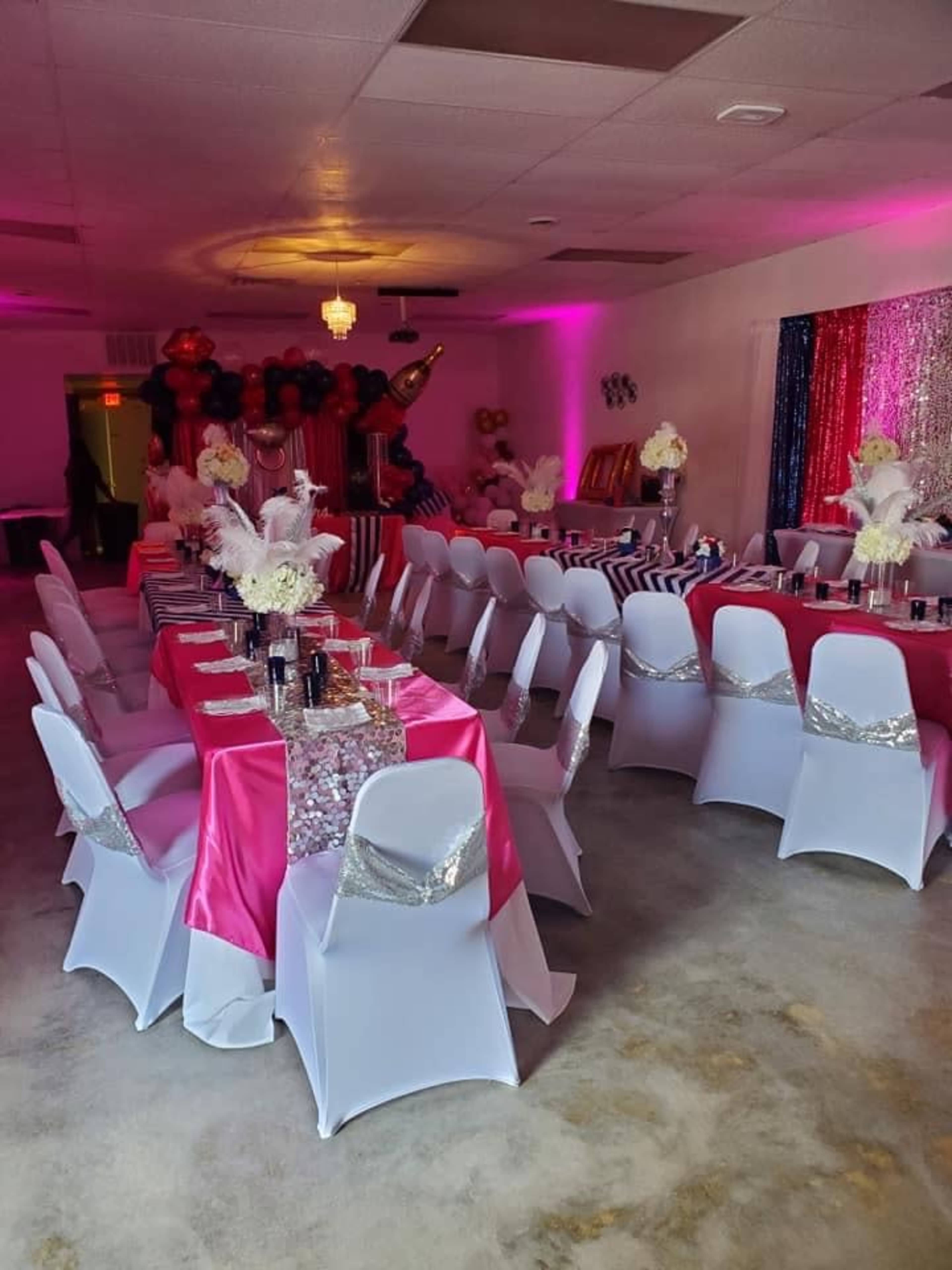 A decorated event space with tables covered in pink and silver tablecloths, arranged with floral centerpieces and set for a celebration.