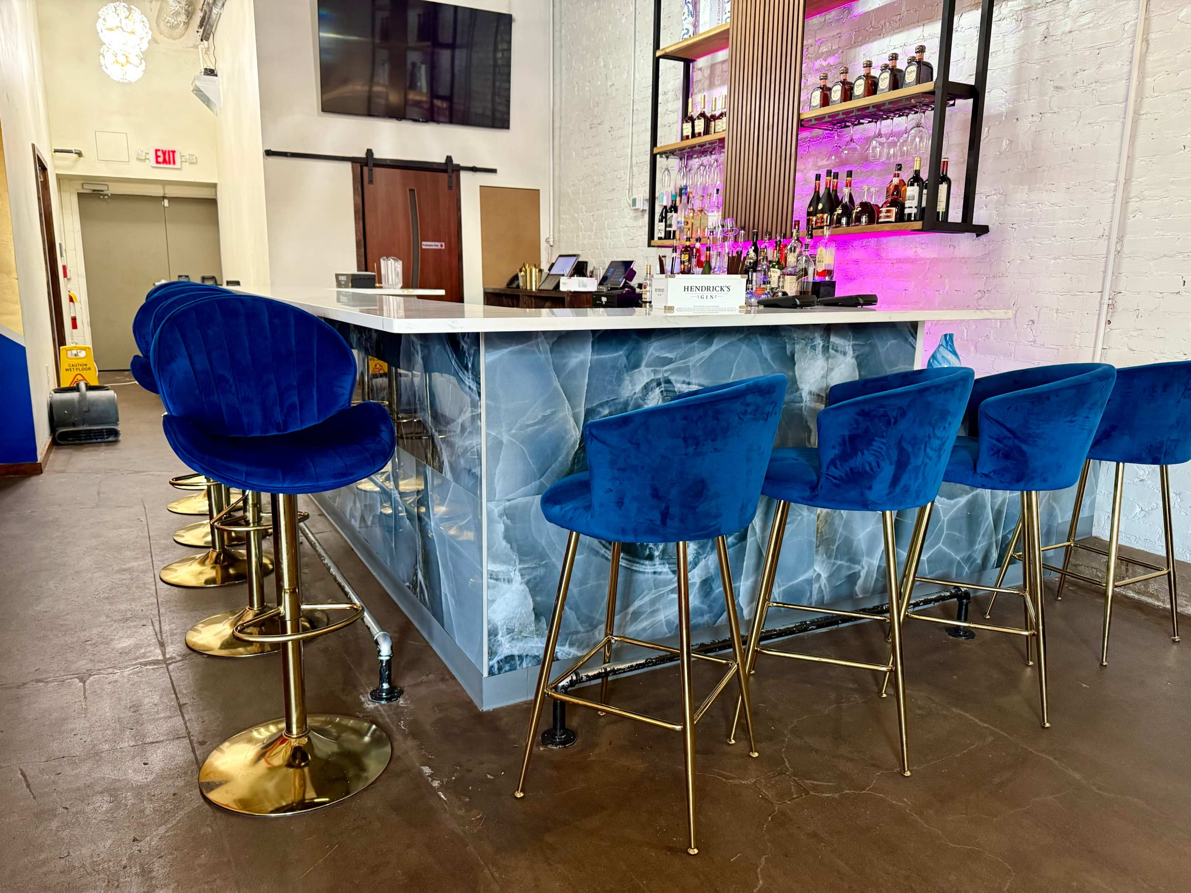 The image shows a modern bar area featuring a marble-patterned counter, blue upholstered chairs, and shelves stocked with various bottles of alcohol against a white brick wall.