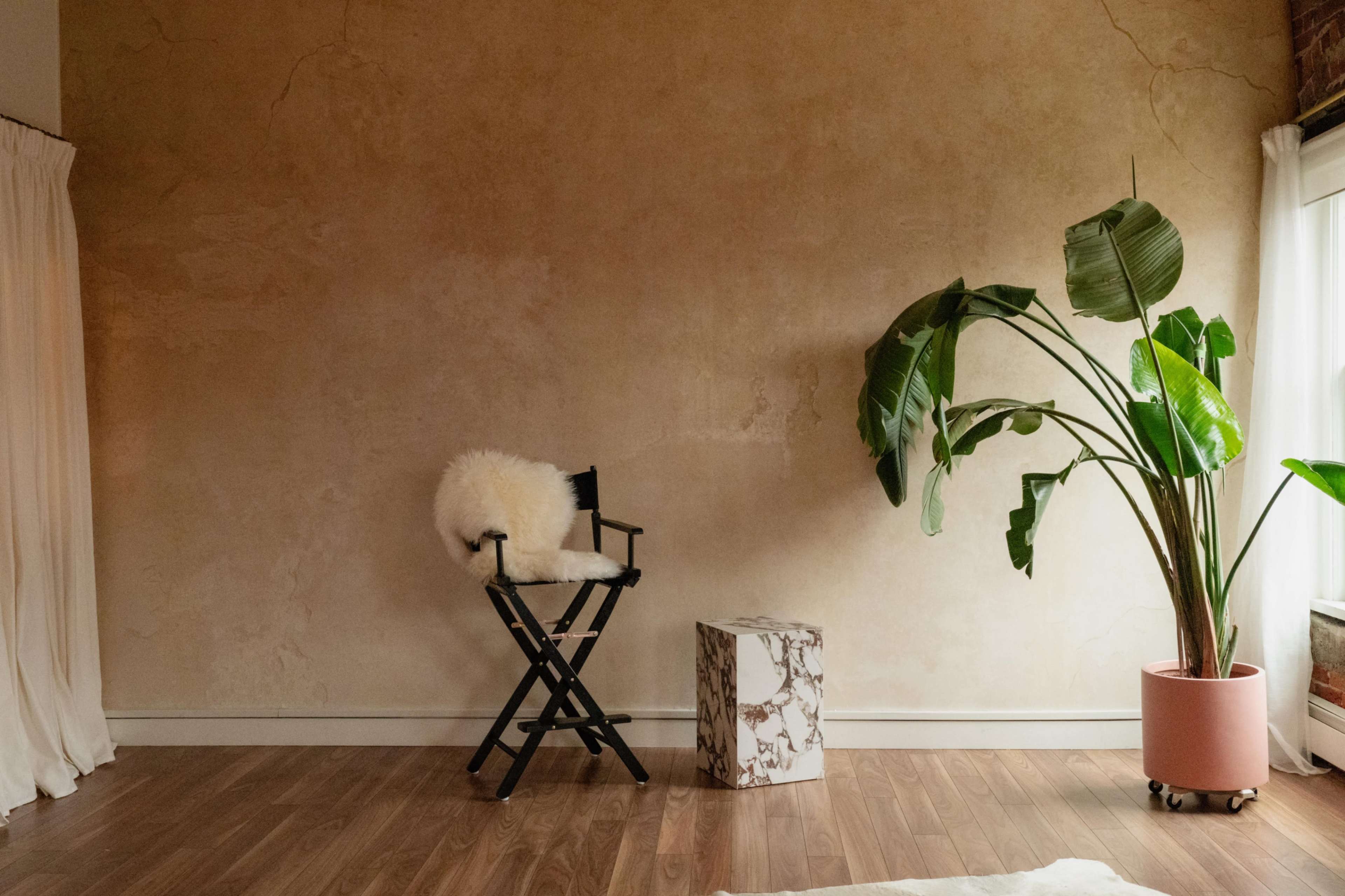The image shows a minimalist interior with a textured beige wall, a black director's chair with a white fur seat cover, a marble-patterned side table, and a large green plant in a pink pot.