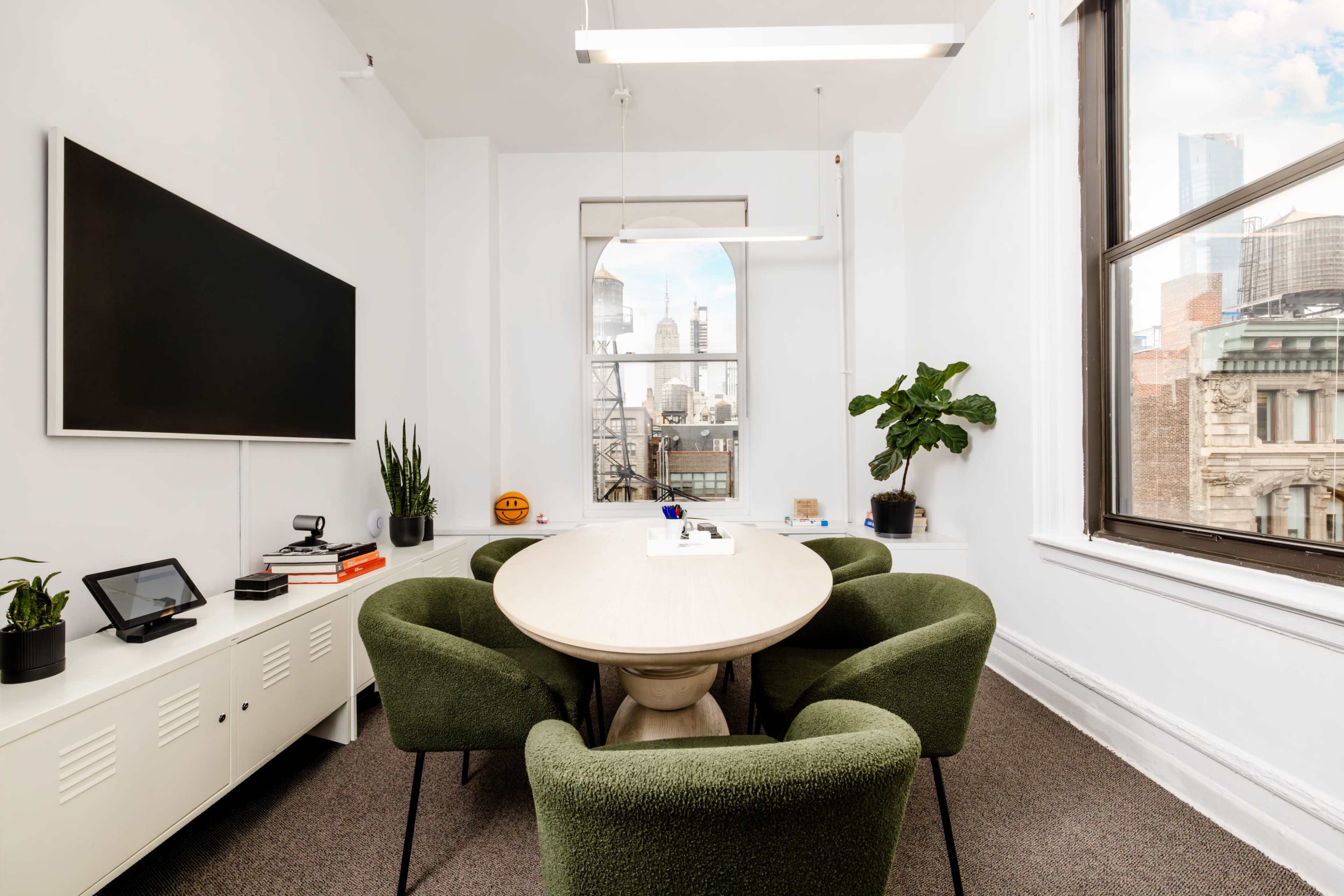 A modern conference room features a round table surrounded by green chairs, with large windows displaying a city skyline and a flat-screen TV mounted on the wall.