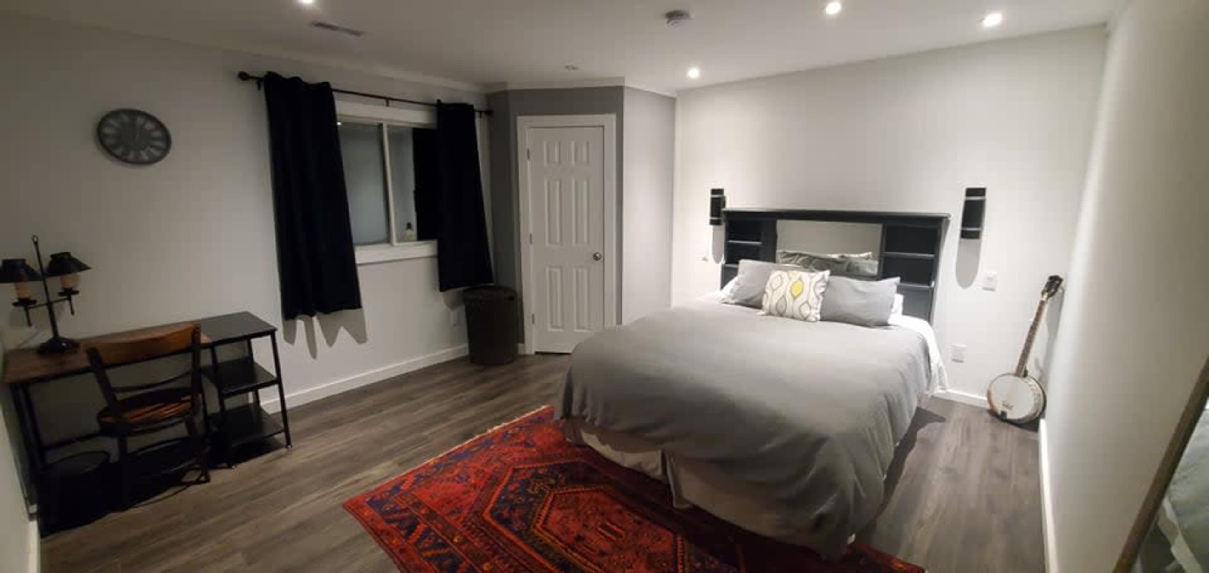 A modern bedroom with a large bed, a desk, a chair, and a patterned area rug on a hardwood floor, surrounded by gray walls and a window with black curtains.