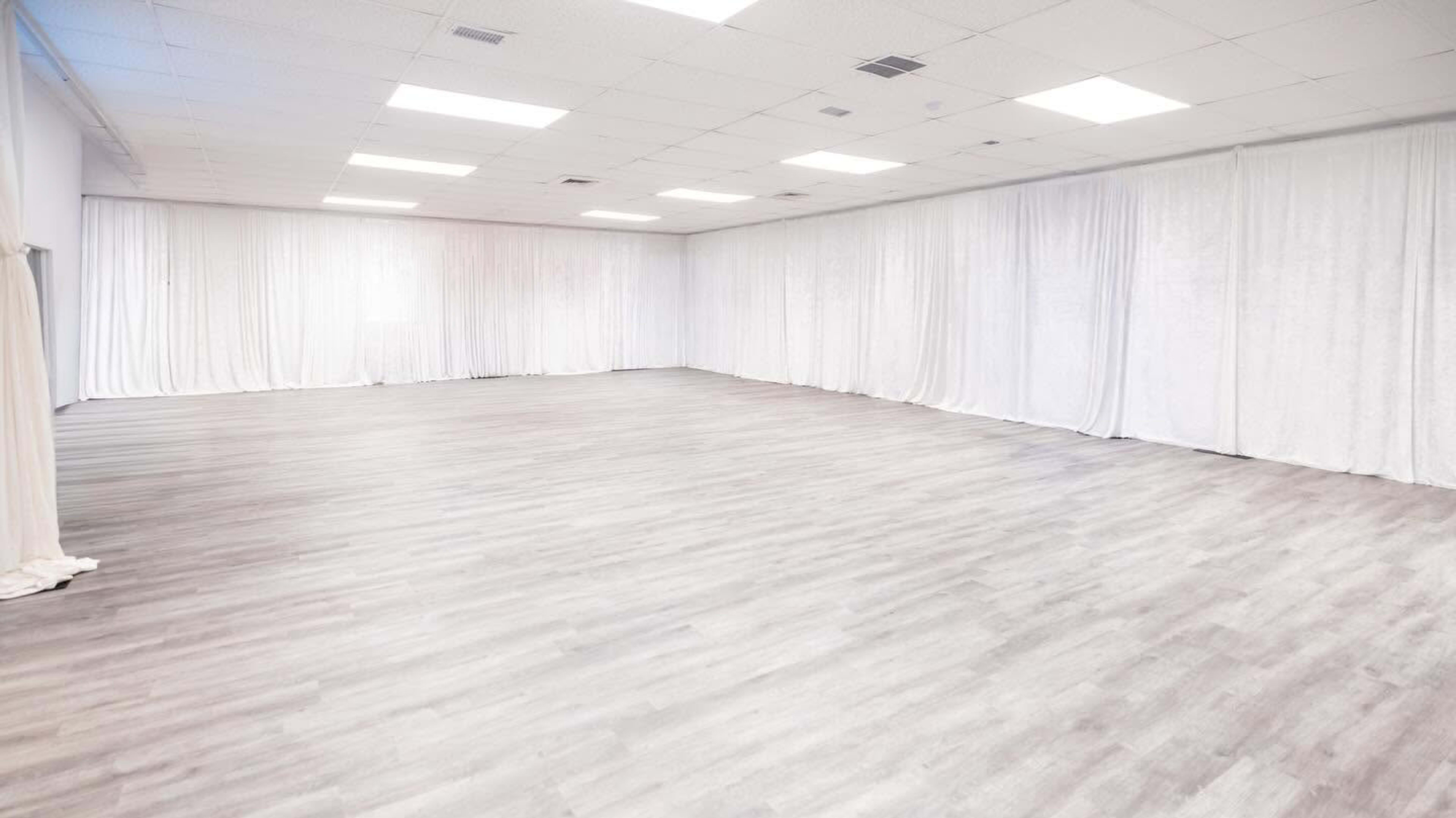 The image shows a spacious indoor room with a wooden floor, white draped curtains, and bright overhead lighting.