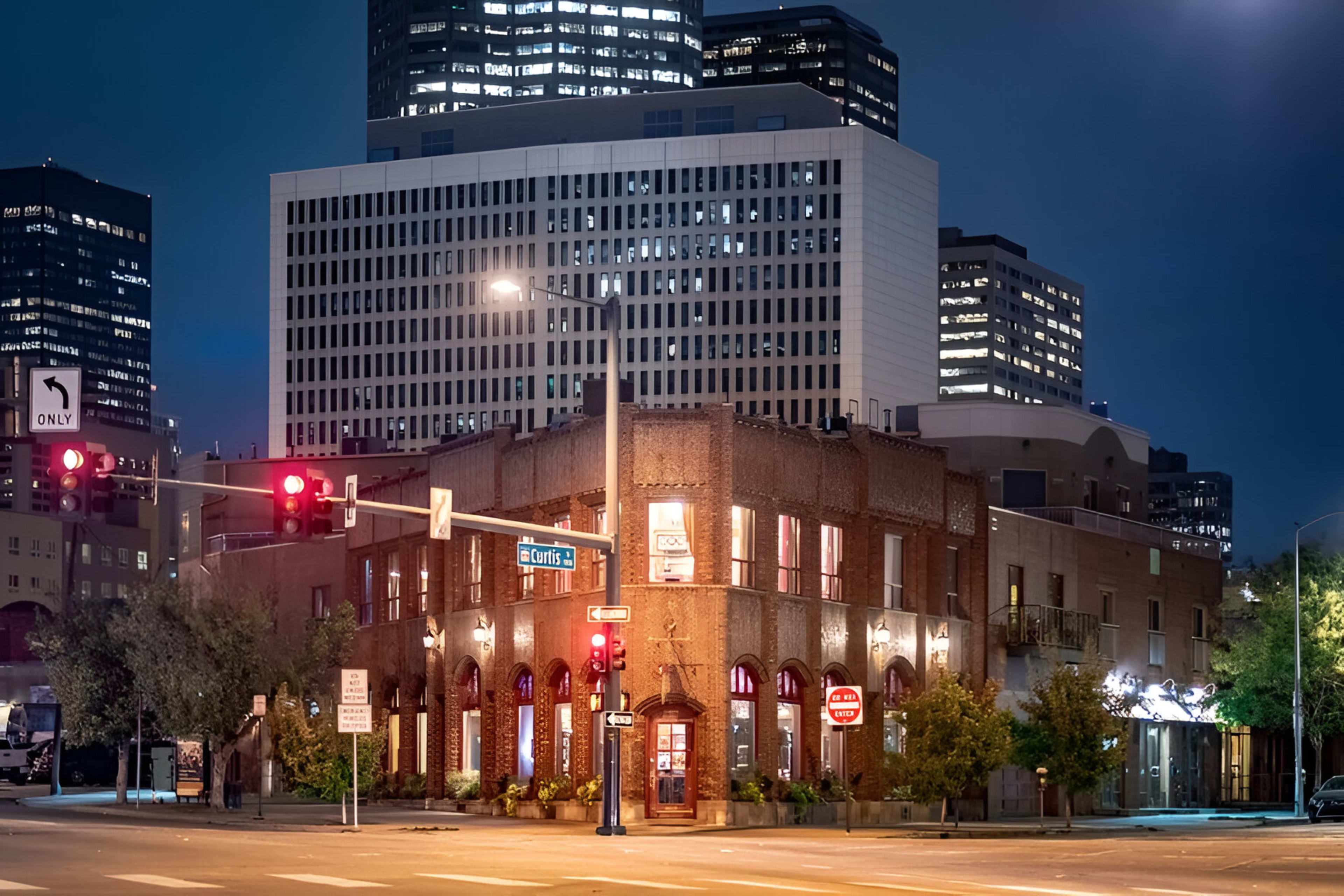 A brick building with large windows stands at an intersection, illuminated by streetlights, with modern skyscrapers in the background.