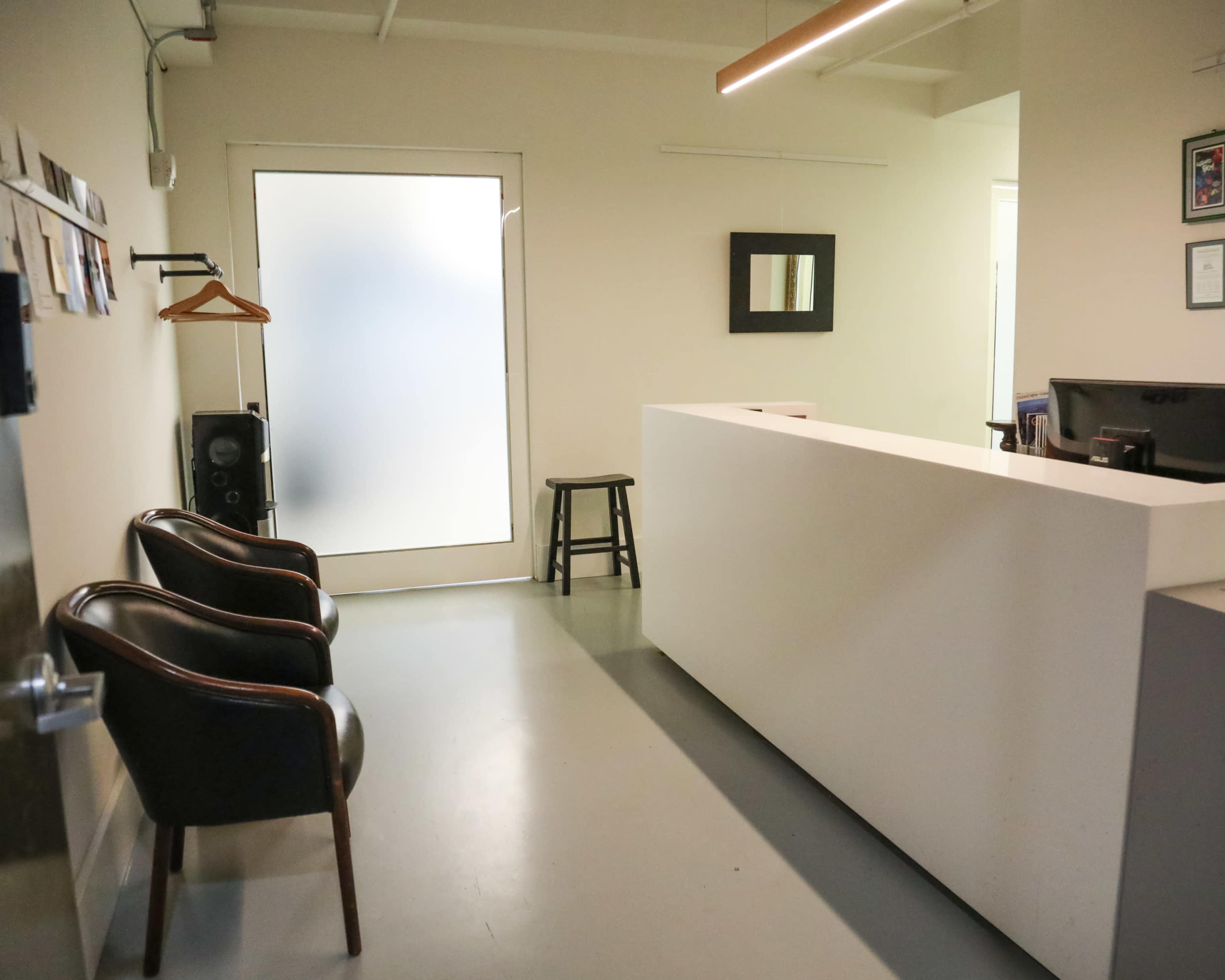 An empty reception area featuring a white counter, two black chairs, a stool, and a frosted glass door.