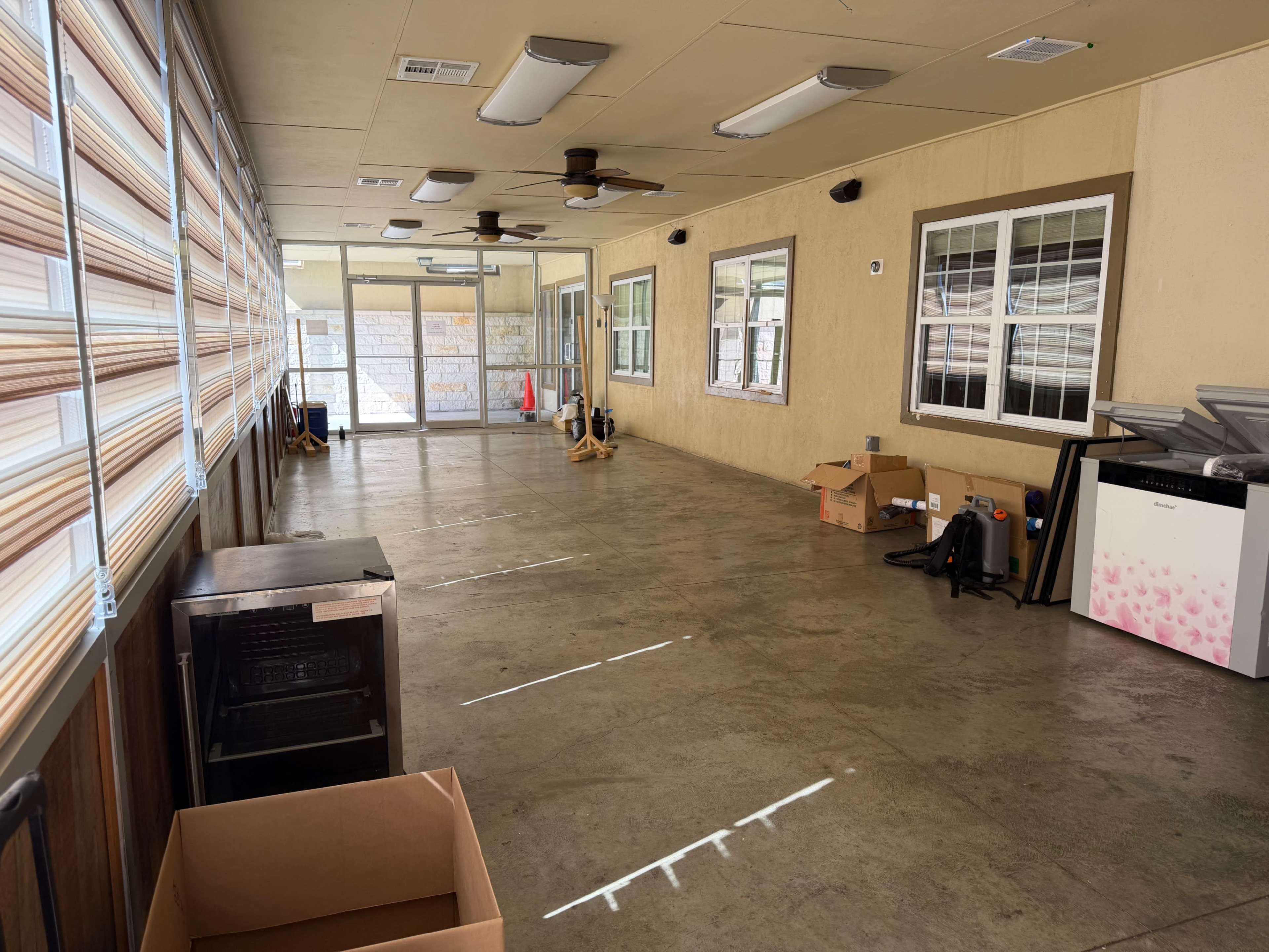 The image depicts a spacious, enclosed room with concrete flooring, large windows, and minimal furnishings, including some cardboard boxes and a white appliance.