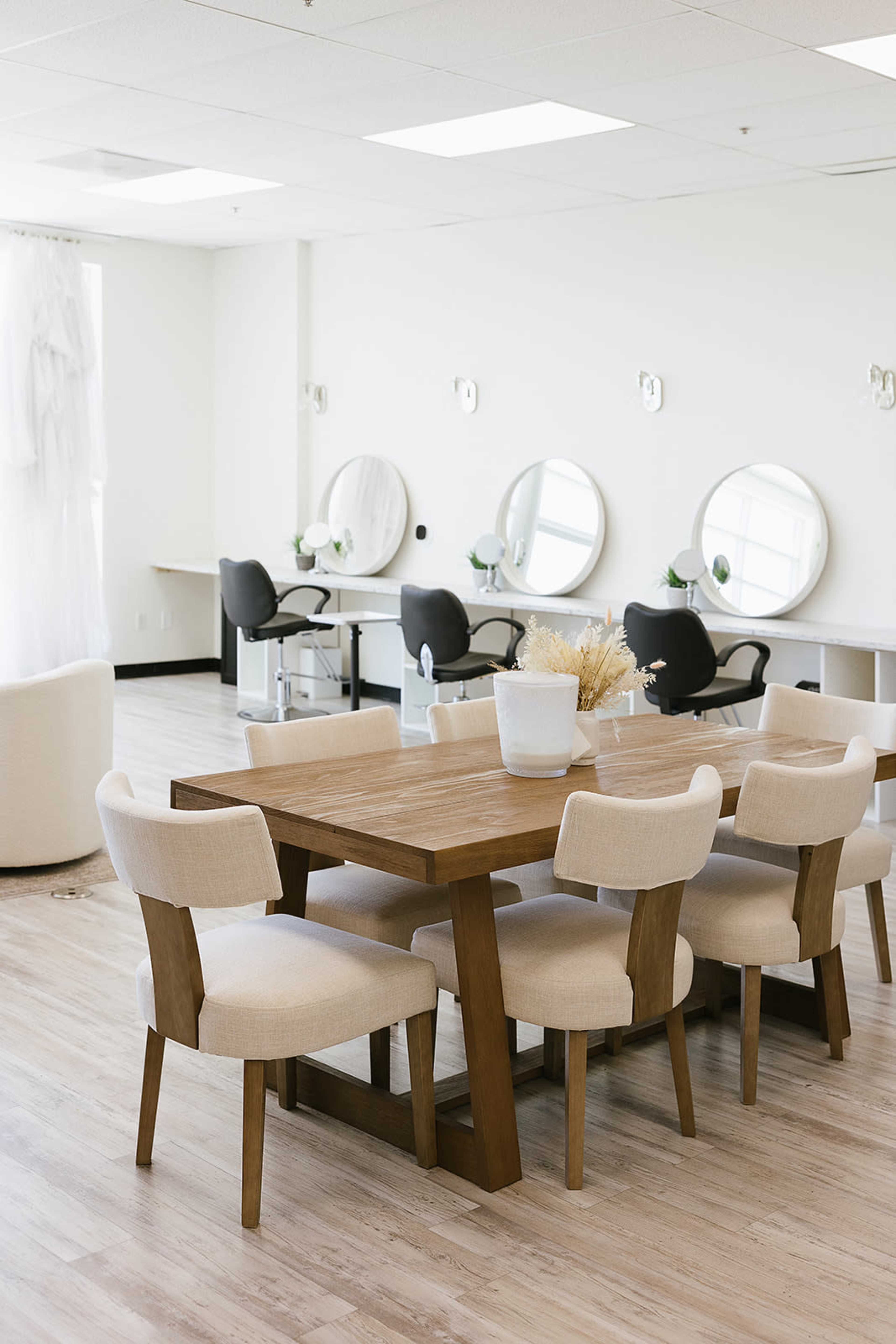The image shows a bright, minimalistic room featuring a wooden dining table surrounded by upholstered chairs, with mirrors and salon chairs positioned along the walls.