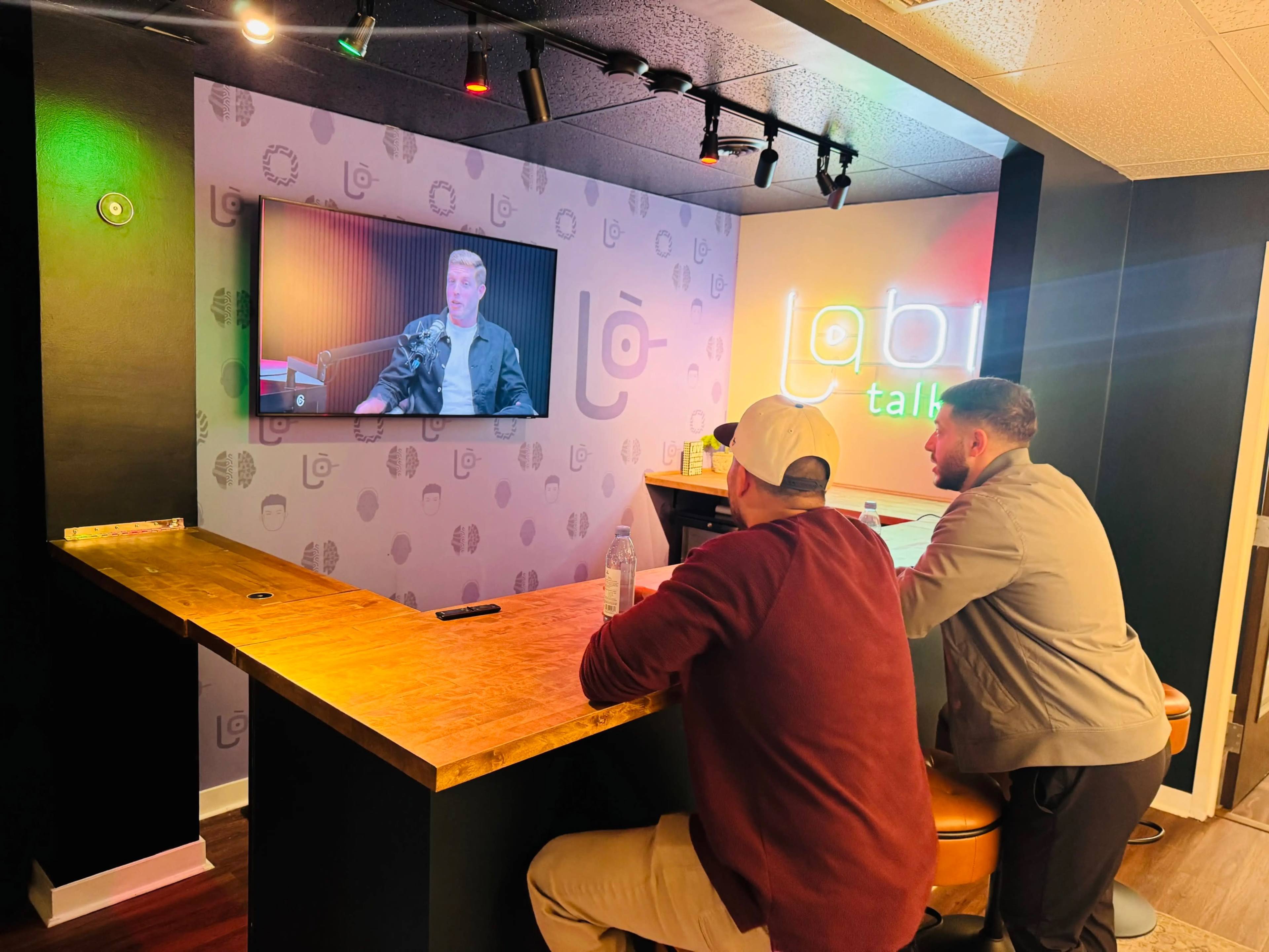 Two men are seated at a wooden bar, watching a man on a television screen in a modern, brightly lit room with a neon sign.