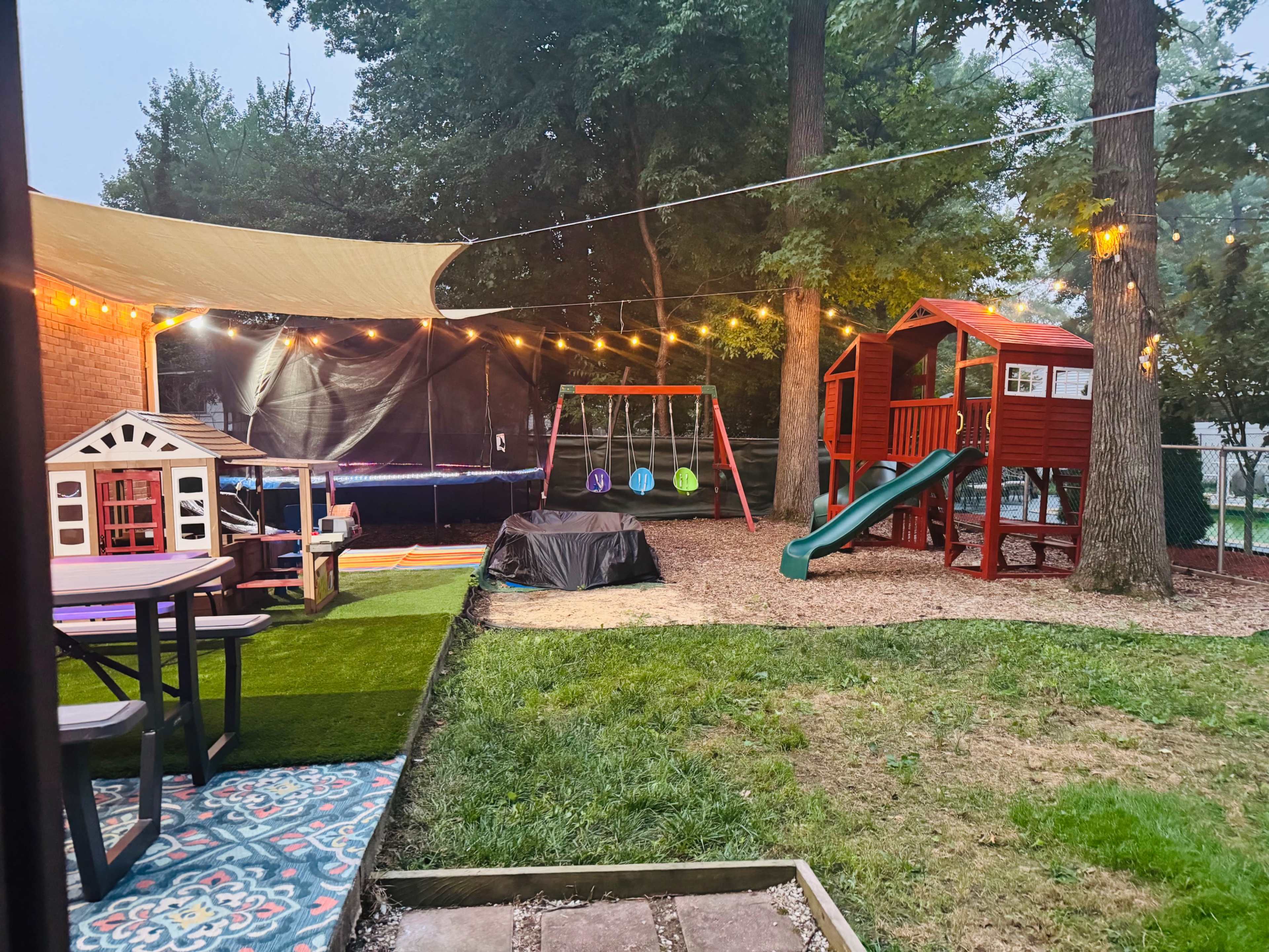 The image shows a backyard playground area with a wooden playset, swings, and a picnic table, all surrounded by trees and illuminated by string lights.