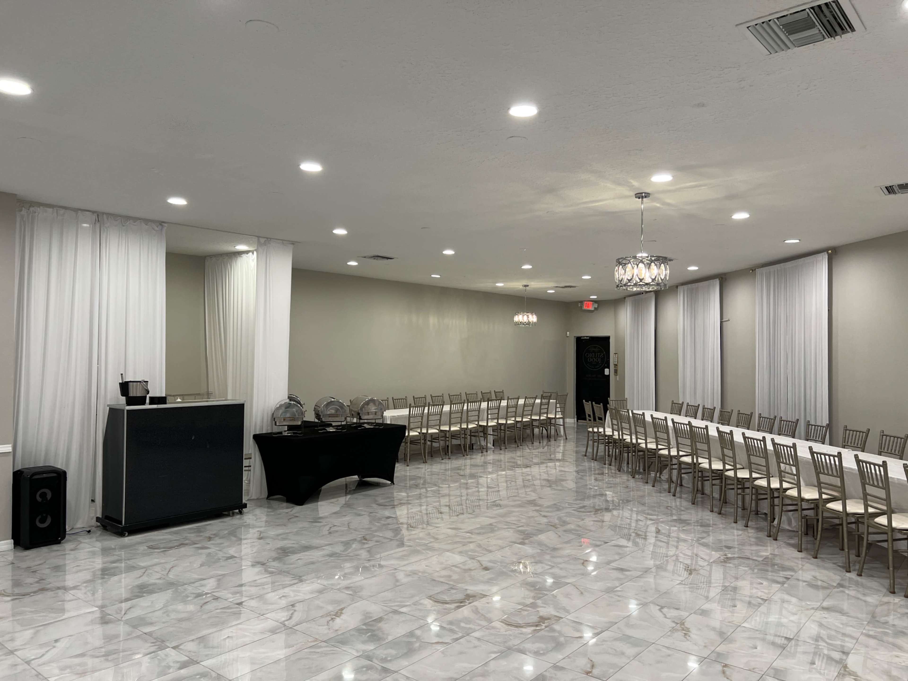 The image shows a spacious event room with marble flooring, long tables set up in a row, and large windows adorned with sheer curtains.