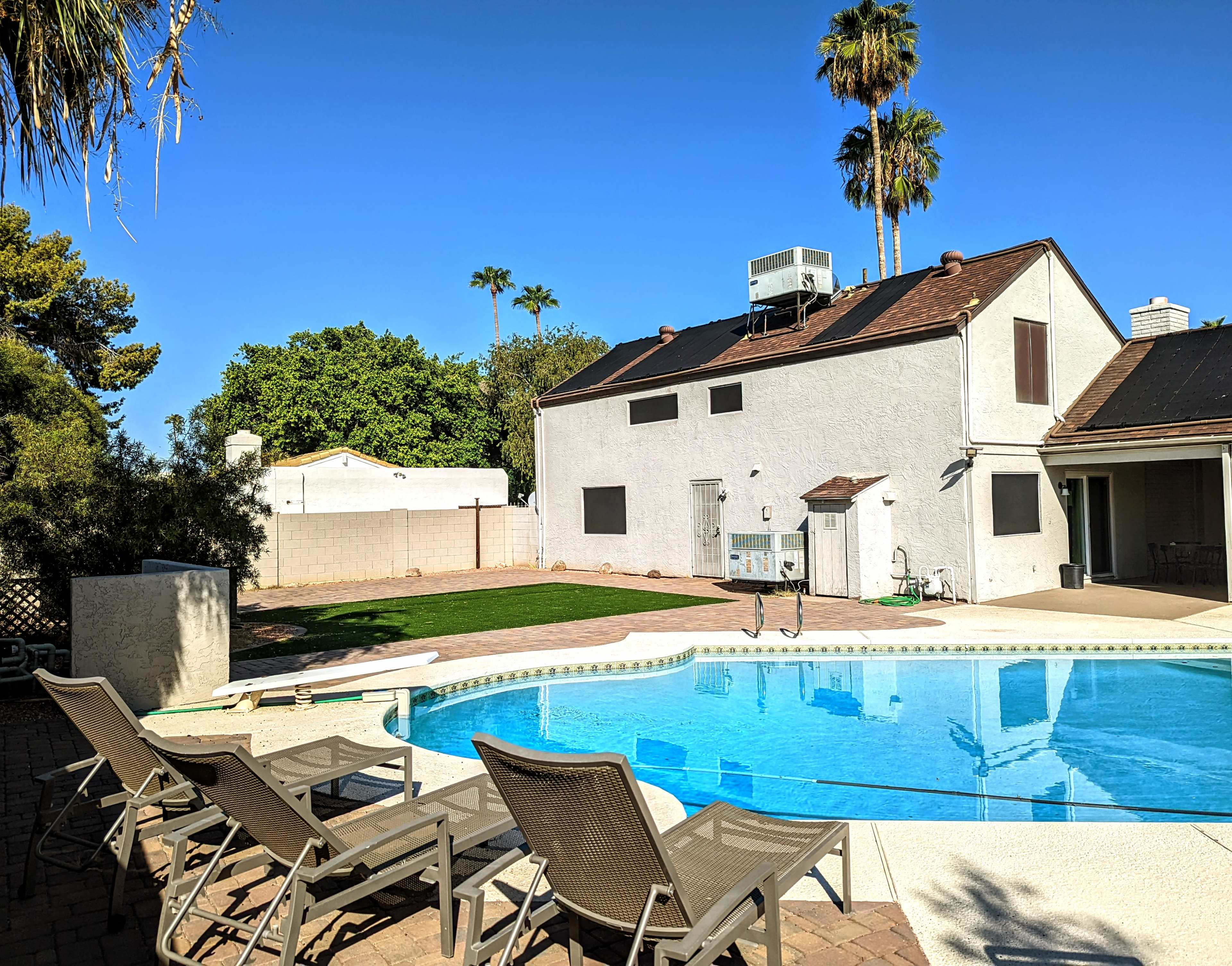 A swimming pool with lounge chairs is situated in the foreground, with a two-story house and palm trees in the background under a clear blue sky.