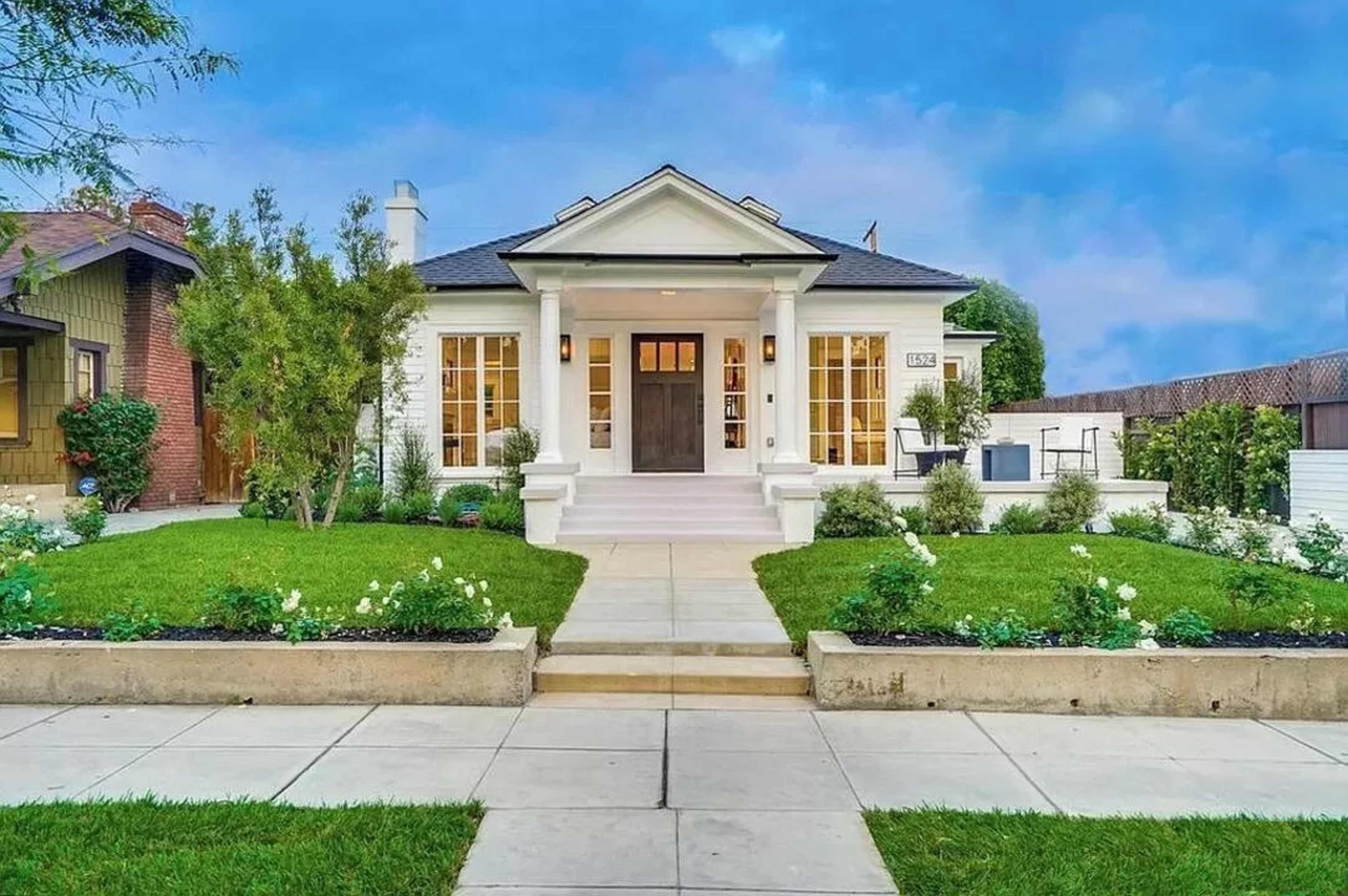 A modern single-story house with a white exterior, a centered entrance, and landscaped grass and flower beds in the front yard.