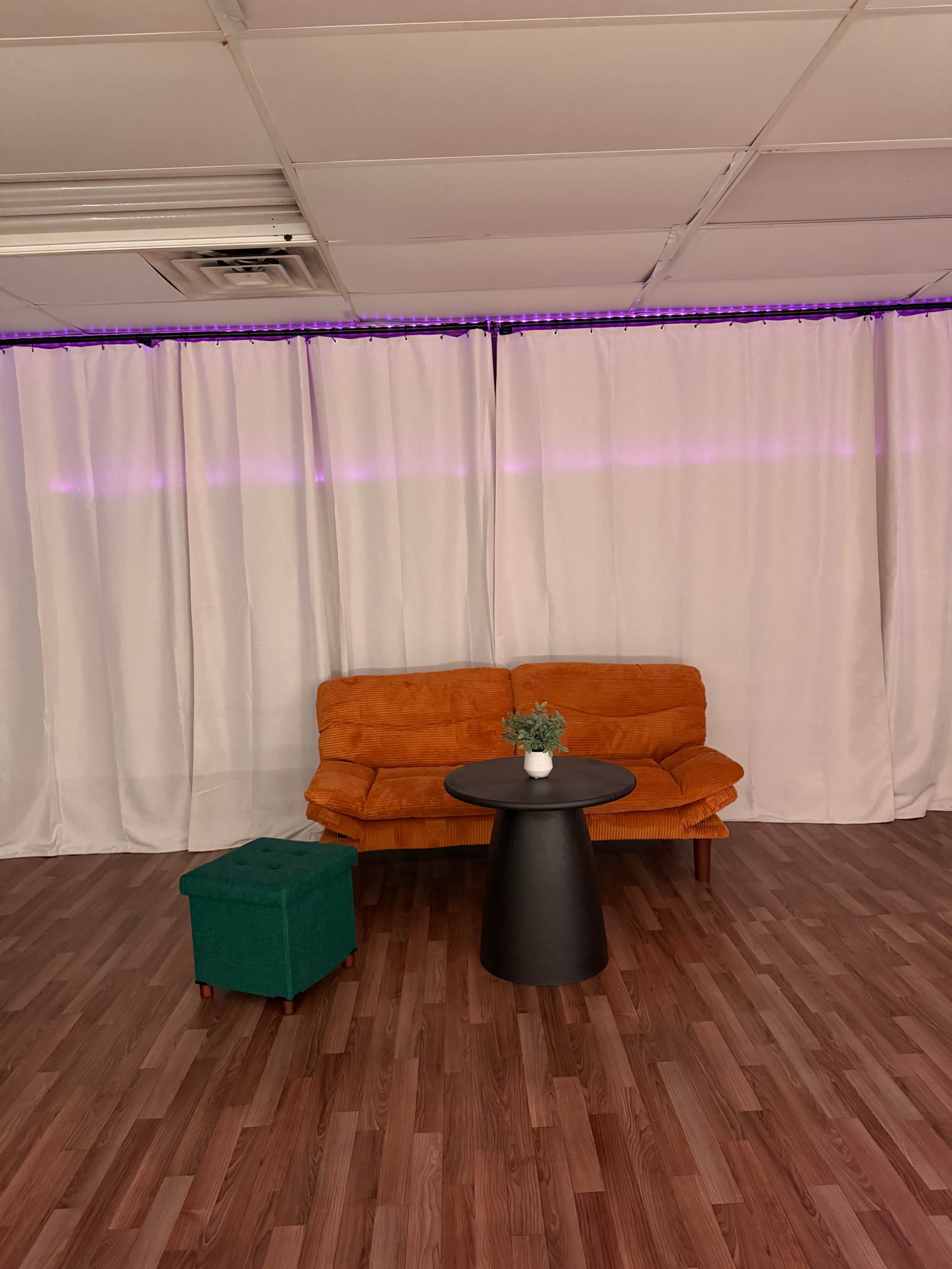 A bright room features an orange couch, a black round table, and a green ottoman, all set against white curtains and wooden flooring.