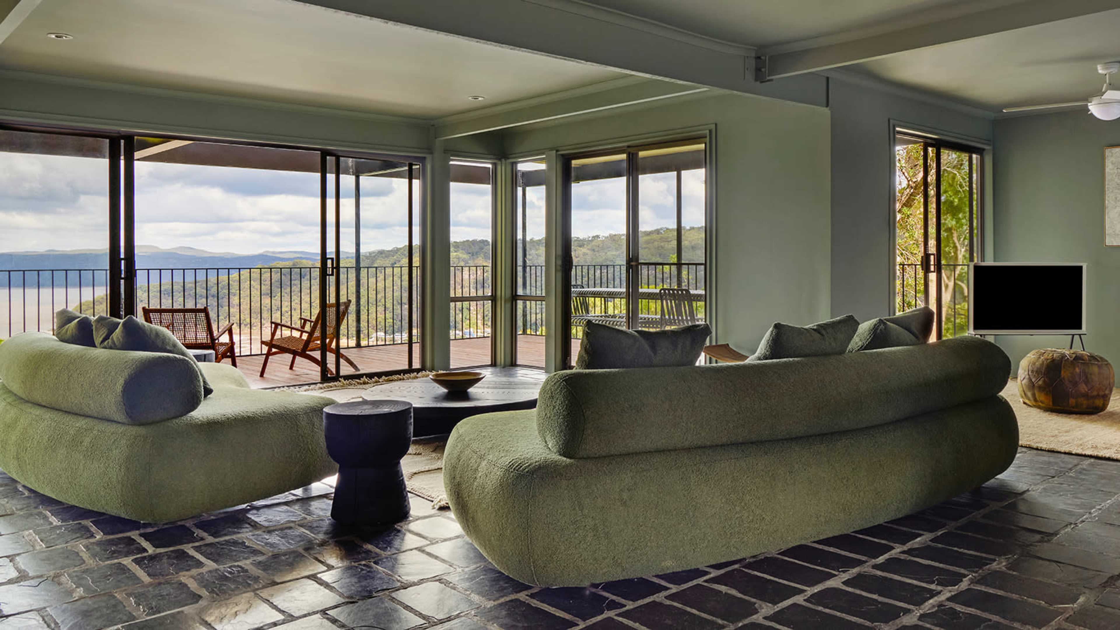 The image shows a modern living room with large windows overlooking a scenic view, featuring green sofas, a black coffee table, and minimalist decor.